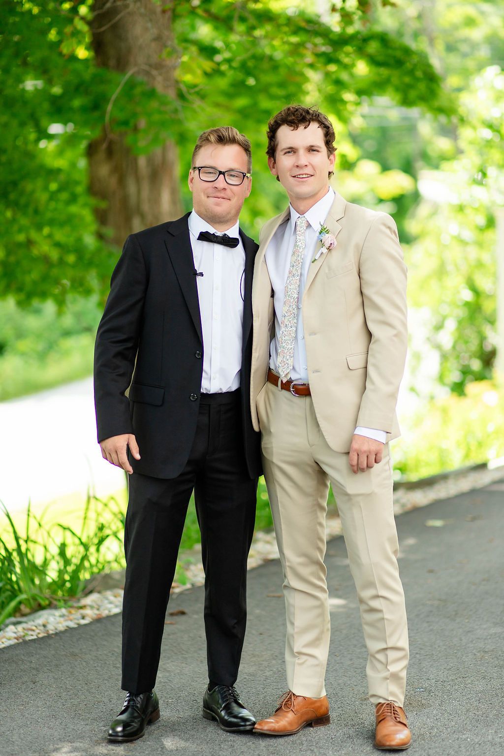 Two men in formal suits smile together outdoors under green trees at a summer wedding celebration.