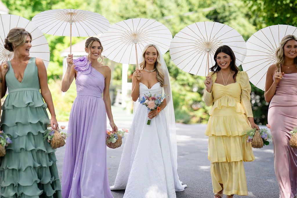 Bride and bridesmaids in pastel dresses holding white parasols while walking outdoors on a sunny day.