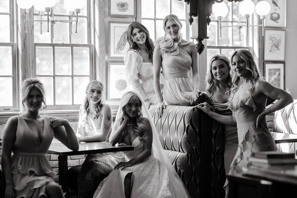 Black and white photo of a bride and bridesmaids posing together in a vintage-style room with large windows.