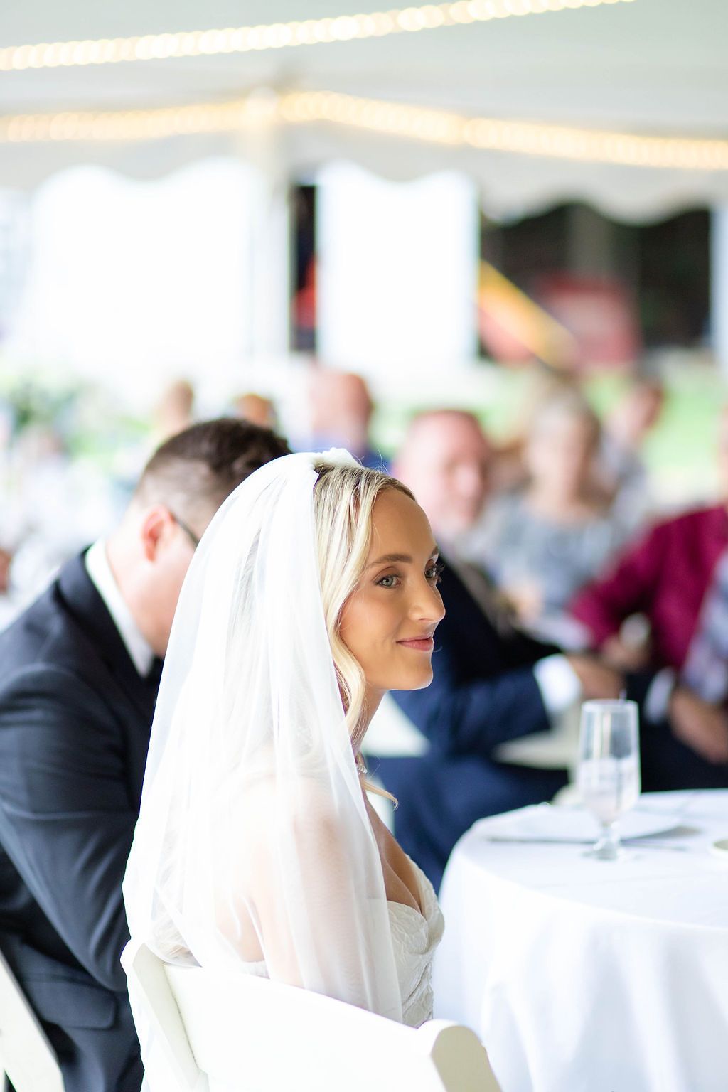 Elegant bride in white veil seated at wedding reception table under a white tent.
