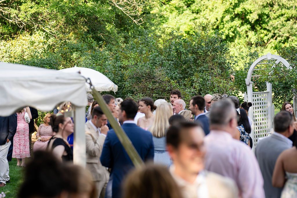Guests gather outdoors at a garden wedding reception beneath lush green trees near a white arbor.