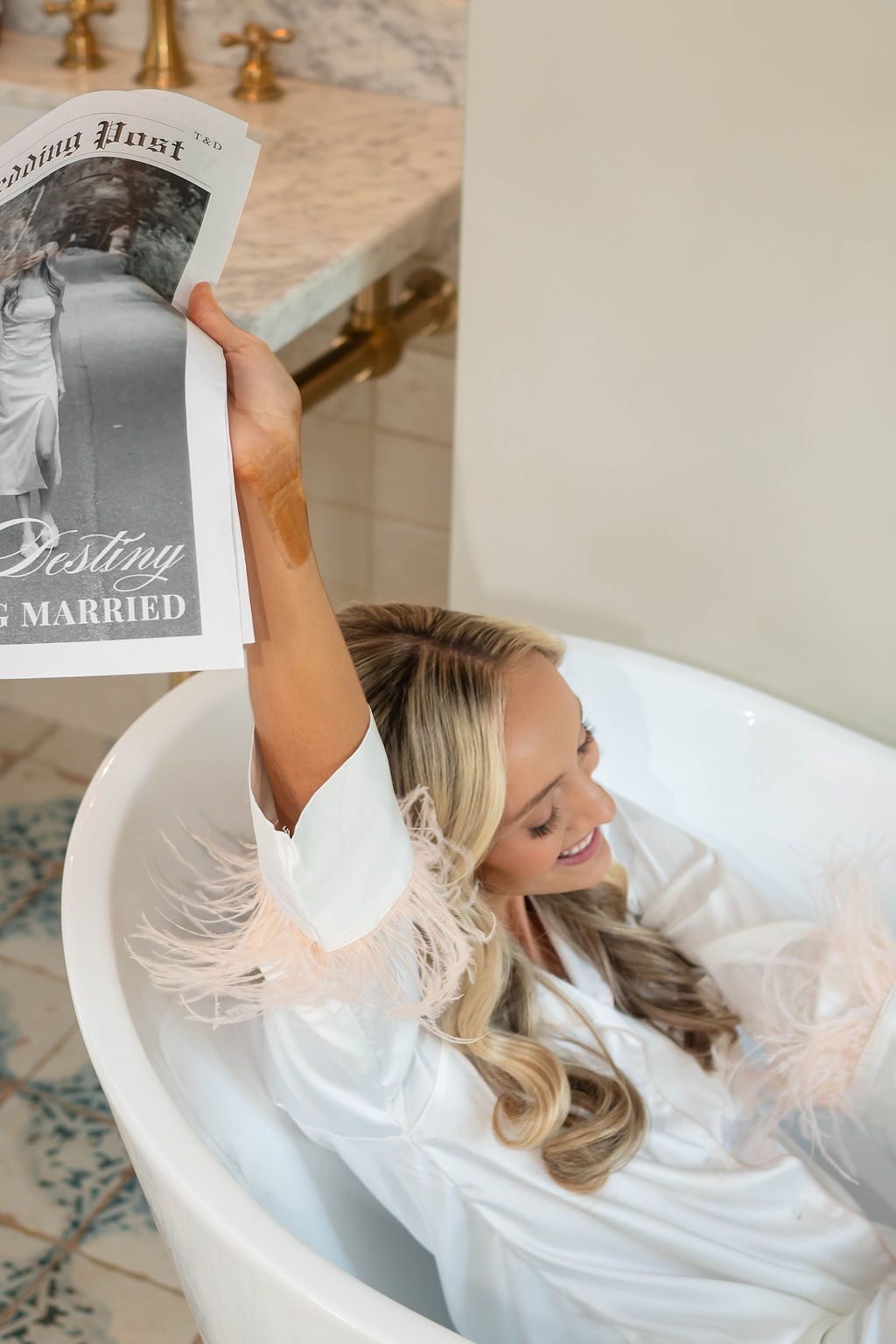 Bride in feathered robe relaxes in bathtub holding personalized wedding newspaper announcement.