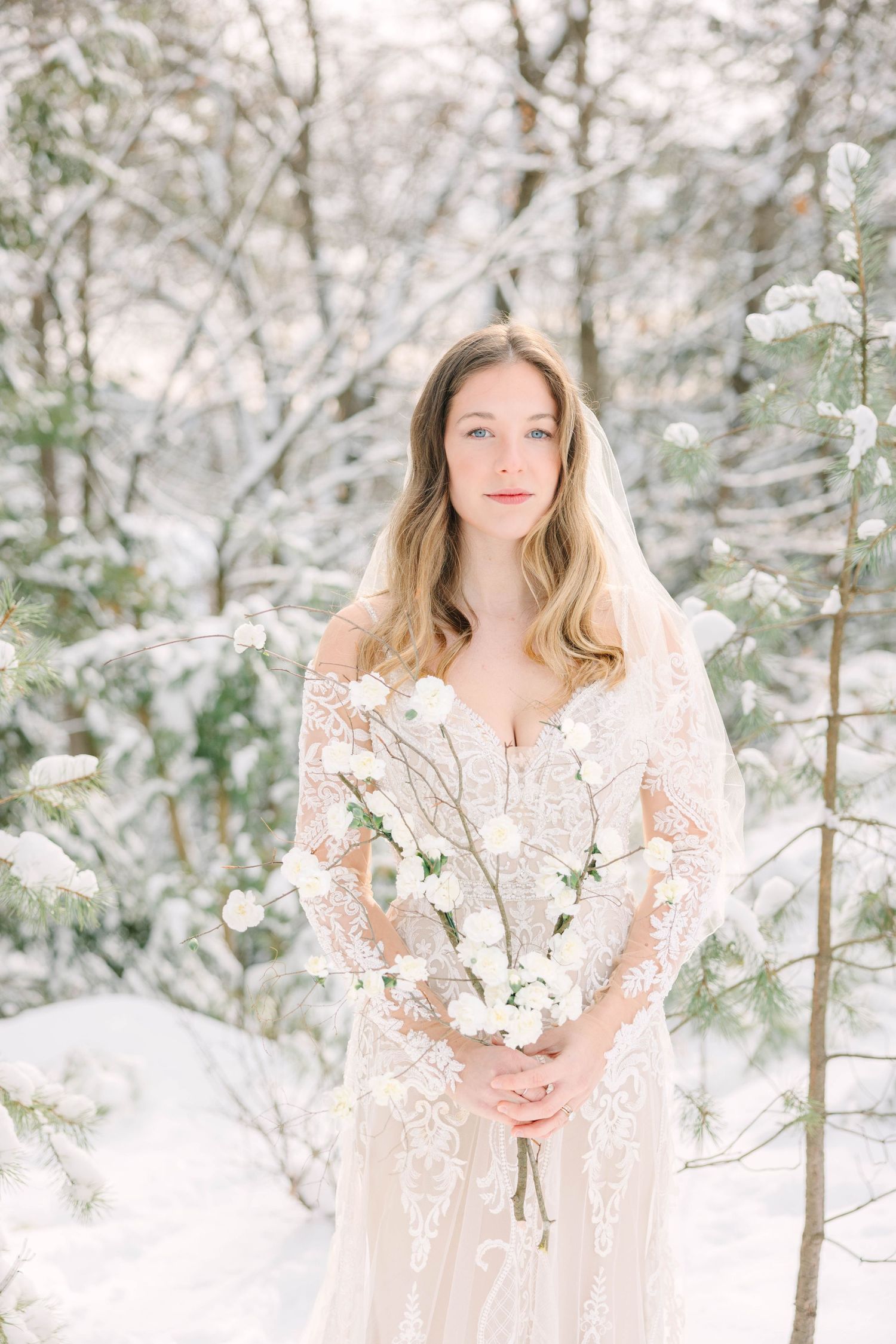 Elegant bride in lace long-sleeve gown holds white blooms in a snow-covered woodland setting.