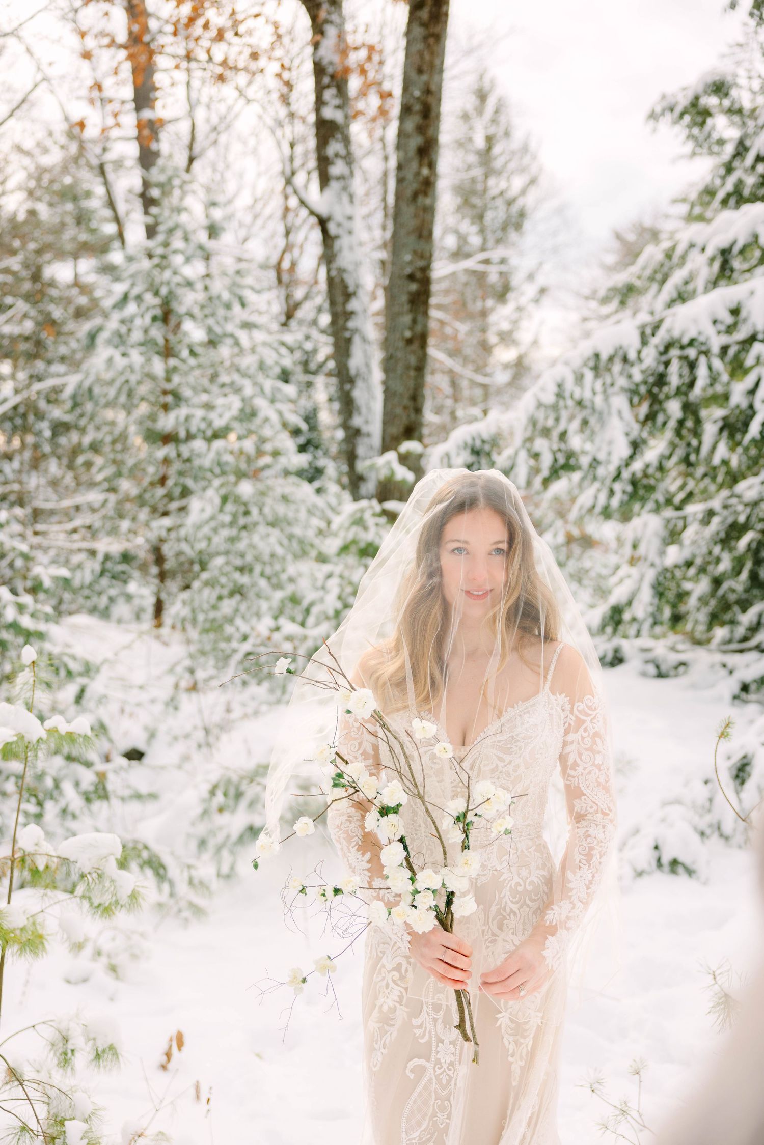 Smiling bride in lace gown and veil holds white bouquet among snow-dusted evergreen trees.