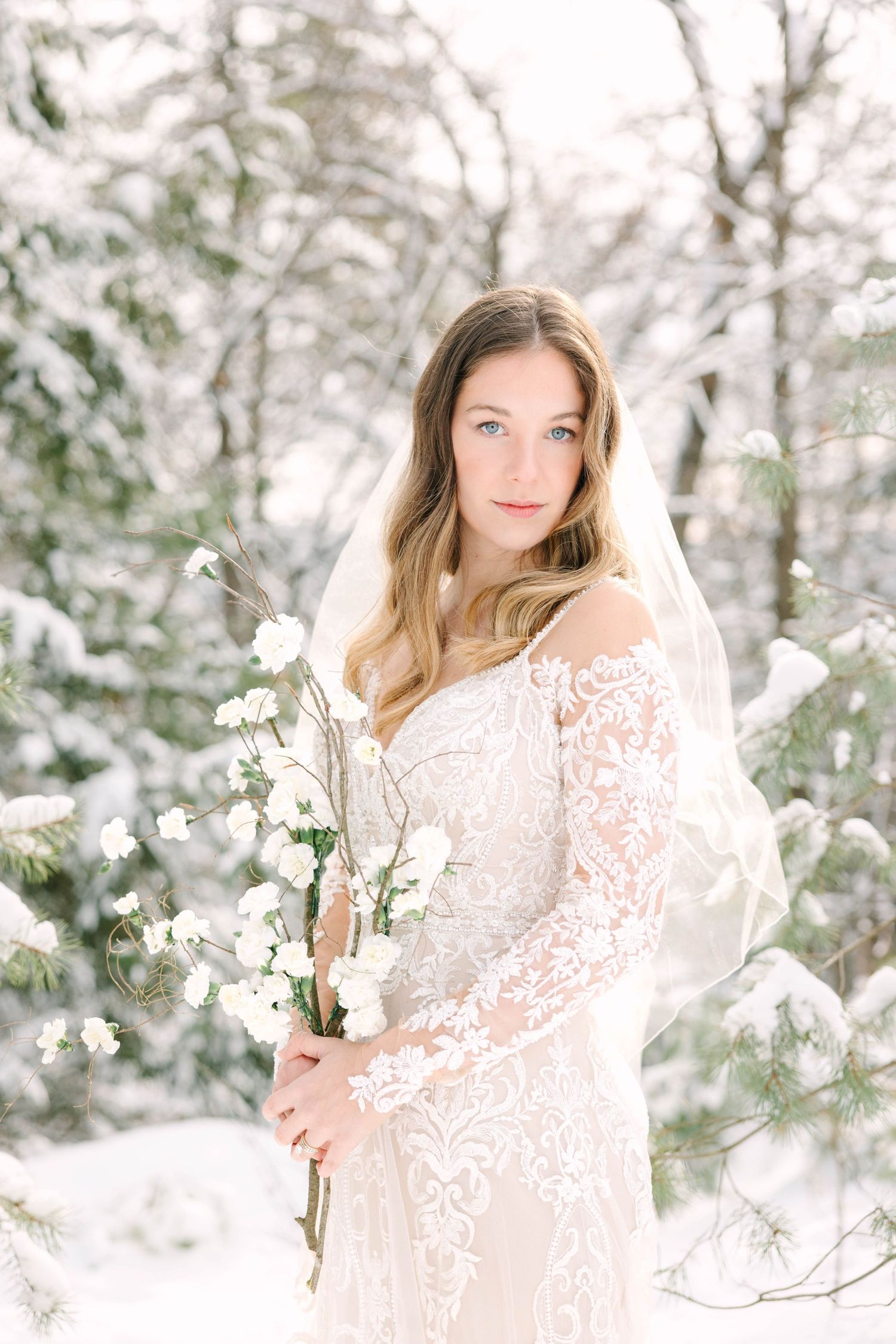 Bride in lace off-shoulder gown and veil holds white bouquet among snow-covered pine trees in winter forest.