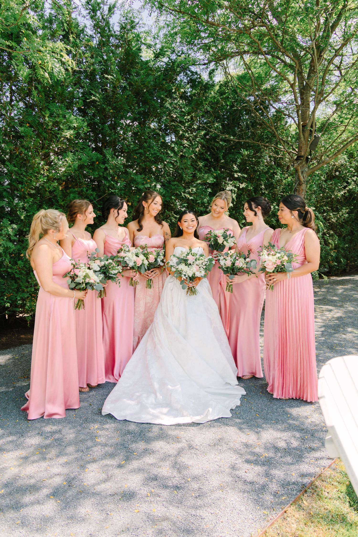 Bride in white gown stands with eight bridesmaids in pink dresses holding floral bouquets outdoors.