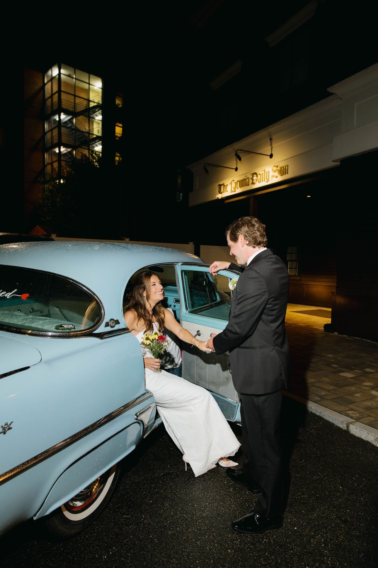 Bride in white gown exits vintage light blue classic car as groom assists her outside evening wedding venue.
