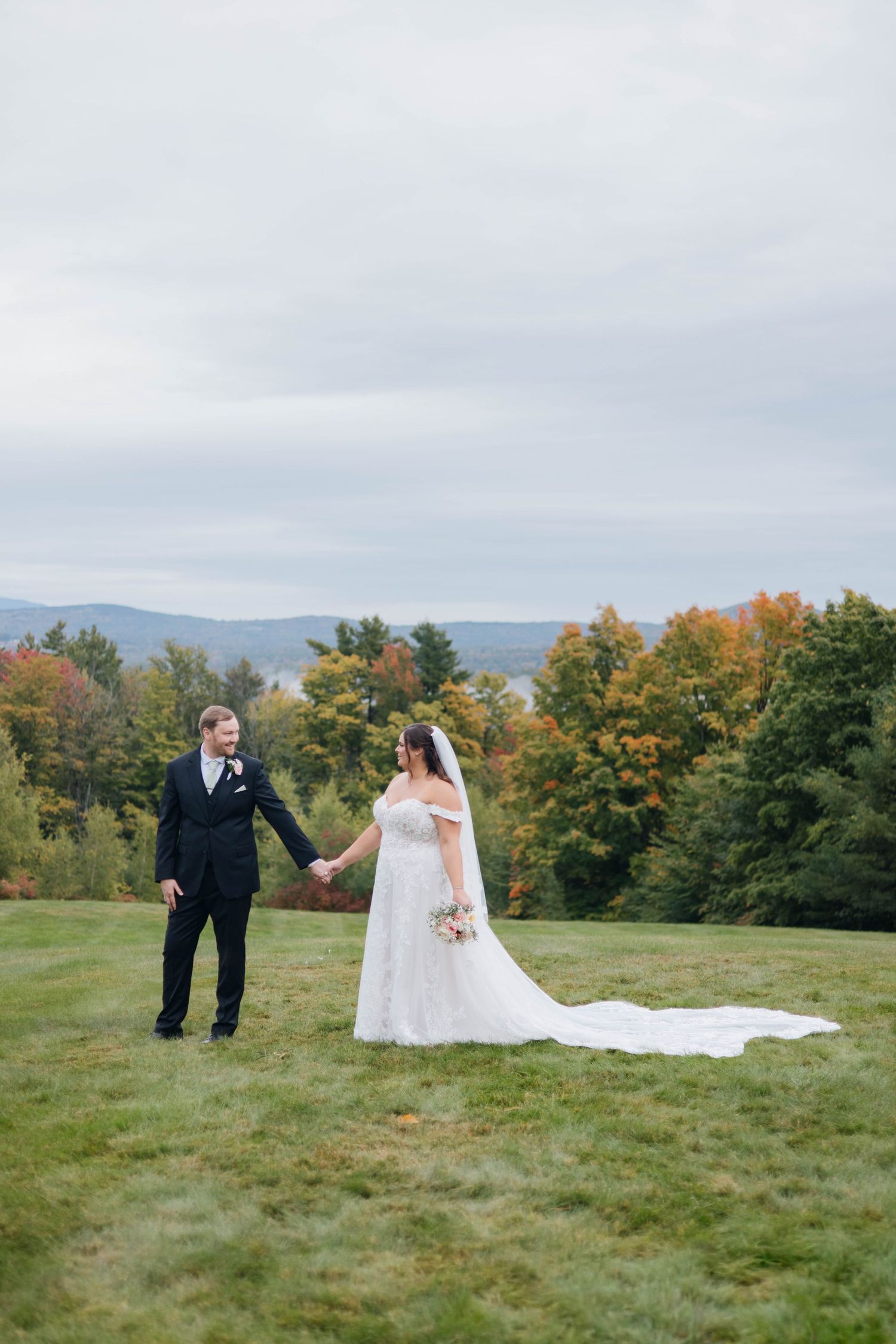 Bride and groom hold hands on a green lawn with colorful autumn foliage and misty mountains in the background.