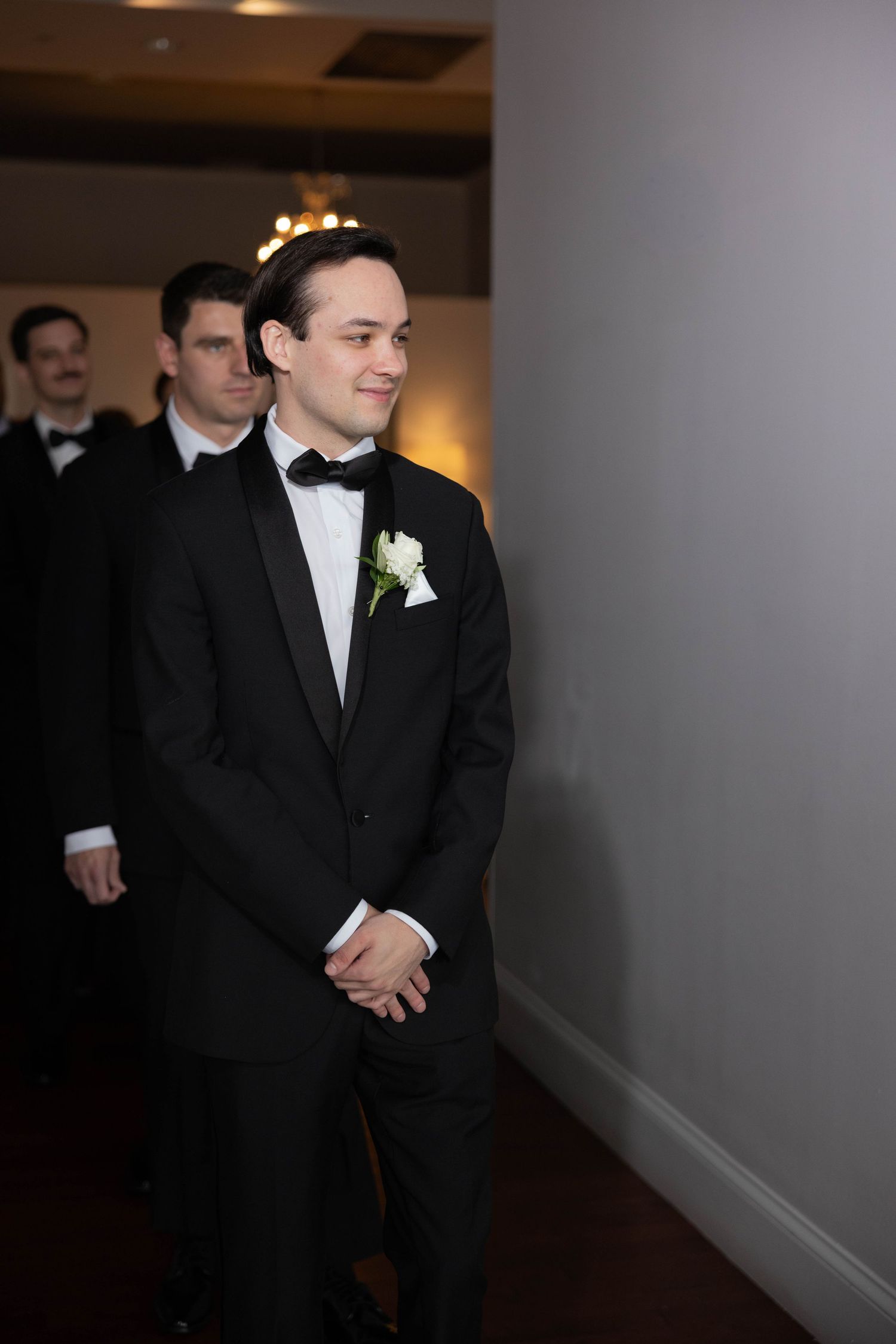 Groom in black tuxedo with white boutonniere smiling while waiting to see bride walk down aisle.