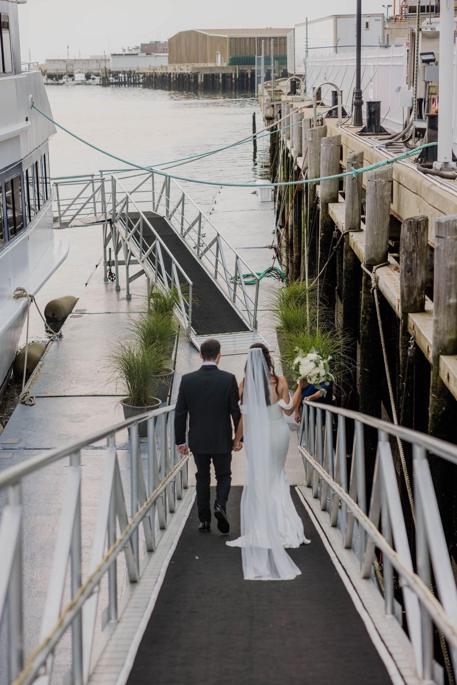 Bride and groom walking along waterfront dock toward marina on their wedding day.