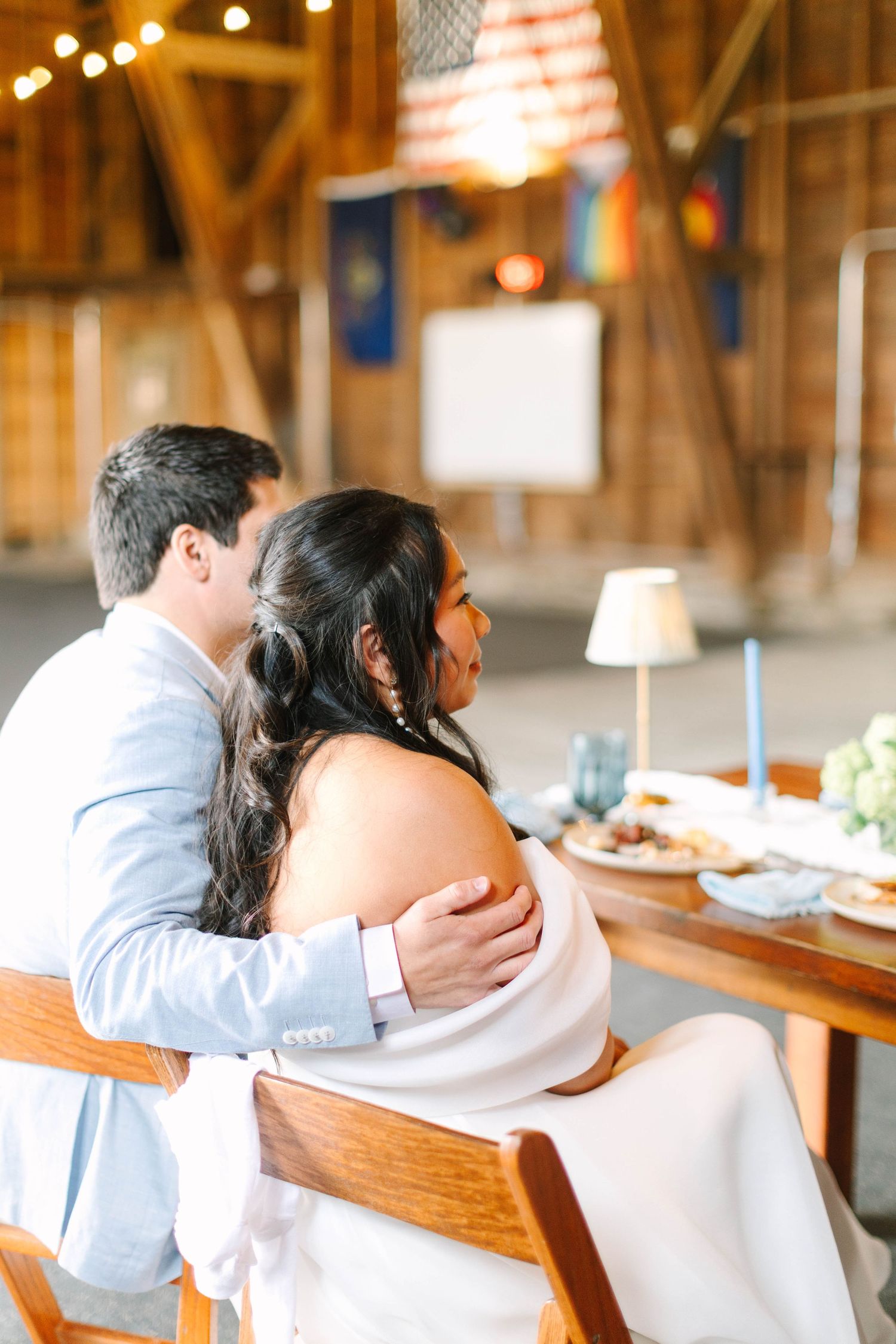 Groom in light blue suit embraces bride in white gown at sweetheart table inside a barn reception.