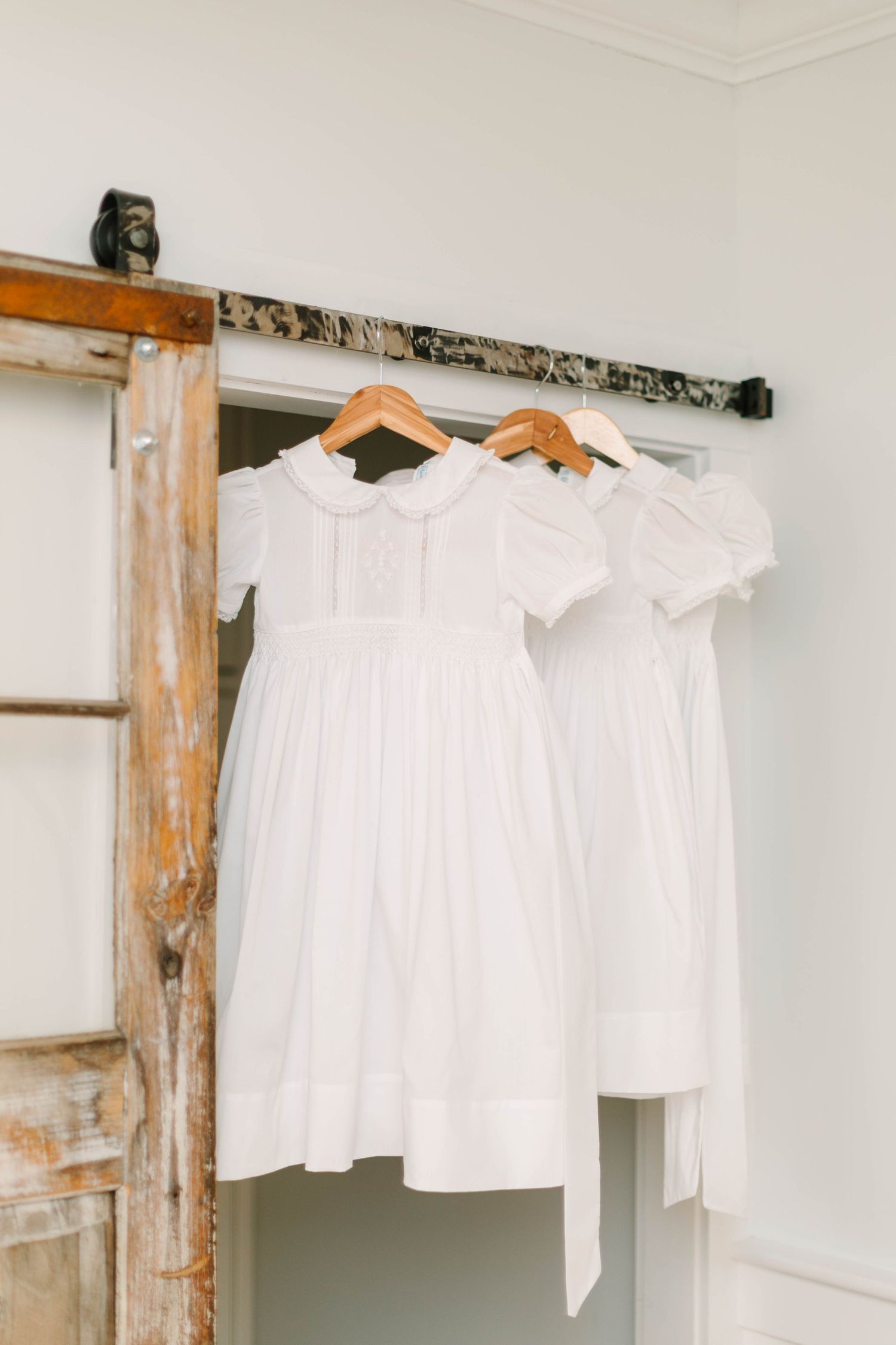 Three white children's dresses hanging on wooden hangers from a rustic barn door rail in a bright white room.