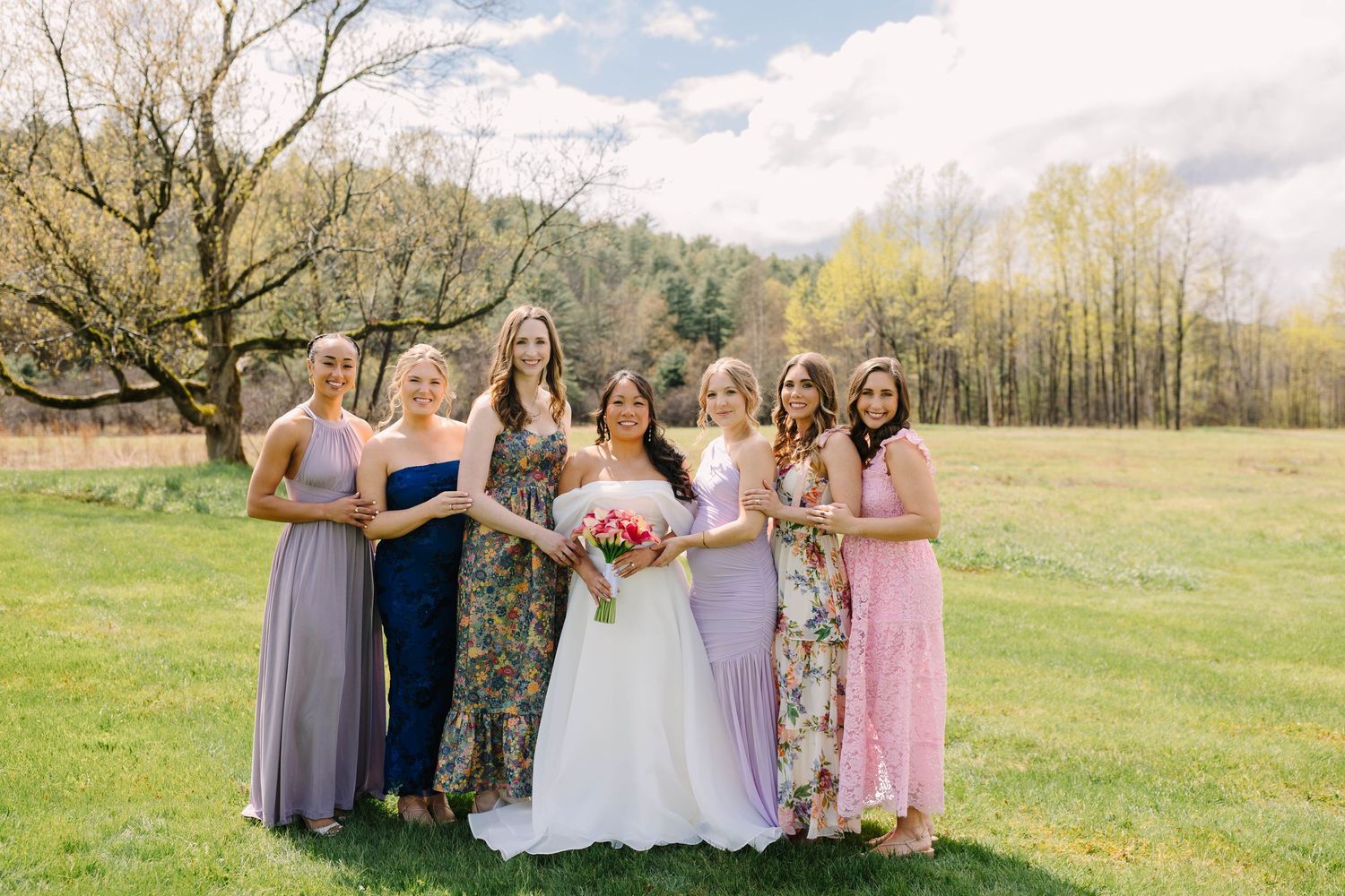 Bride in white gown poses with six bridesmaids in mixed floral and purple dresses on green outdoor lawn.