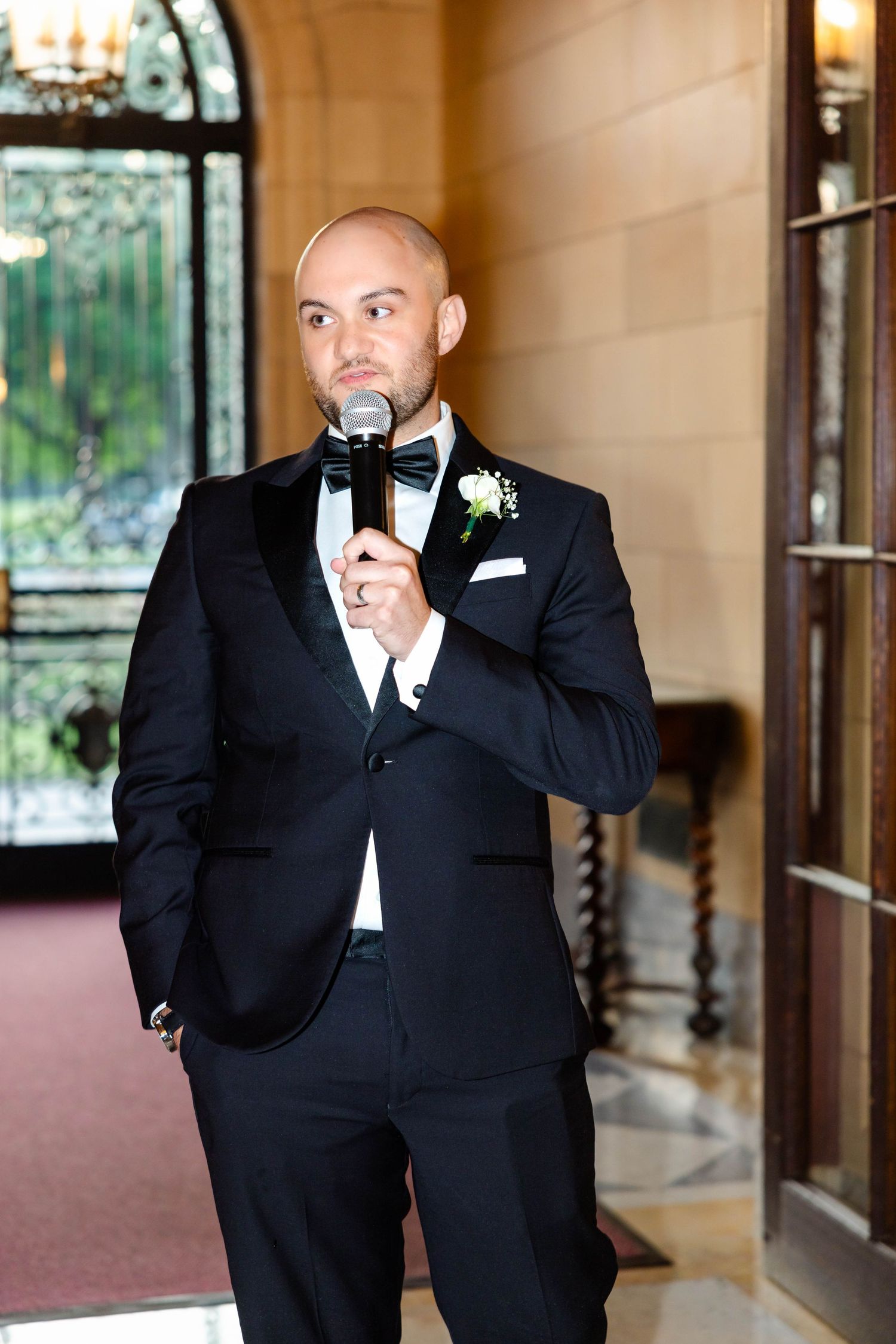 Bald man in black tuxedo speaking into microphone at elegant wedding reception.