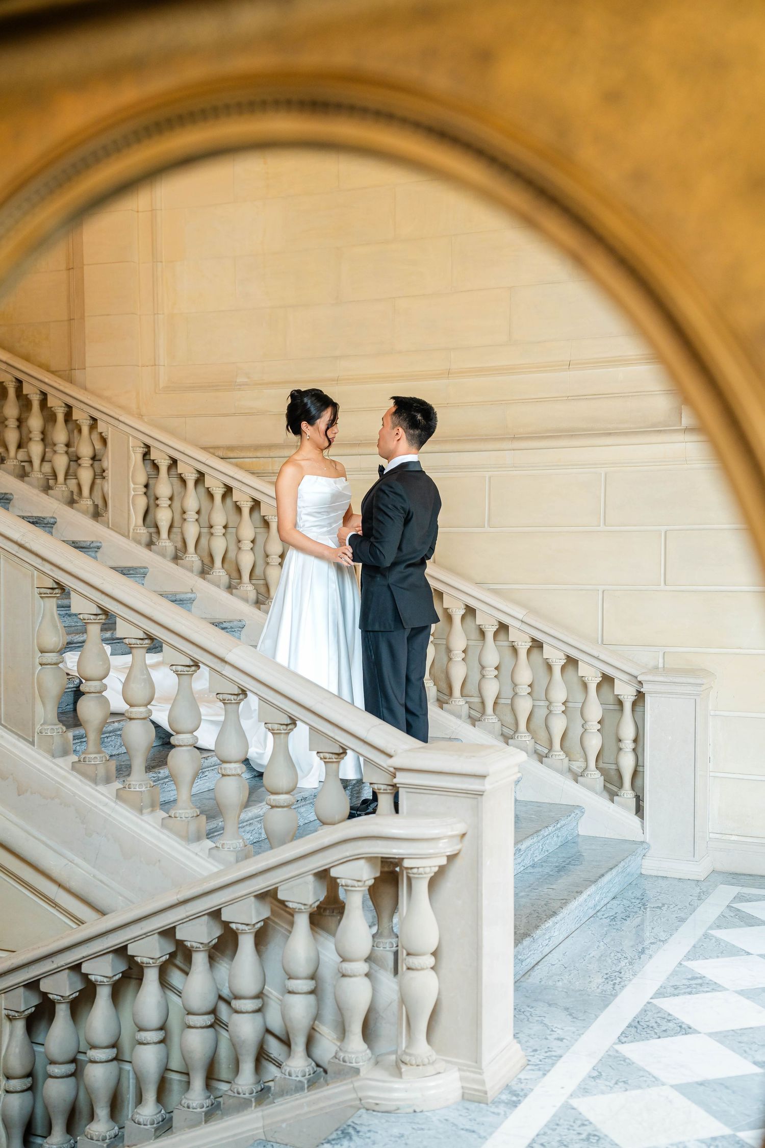 Asian bride and groom hold hands on elegant marble staircase framed by ornate stone archway in grand venue.