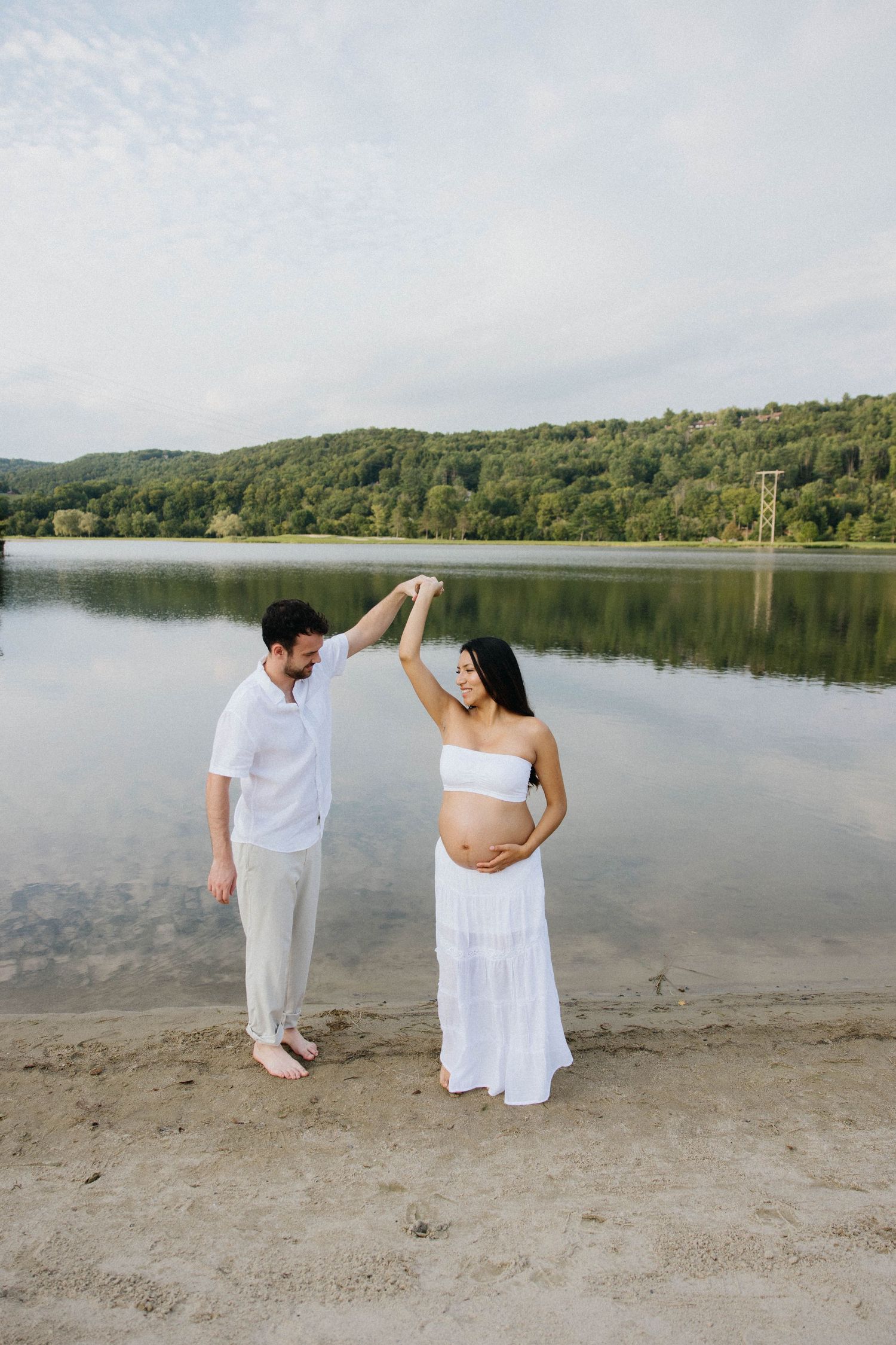 Expecting couple dances by a serene lake, woman in white maternity dress holding her belly while partner twirls her.