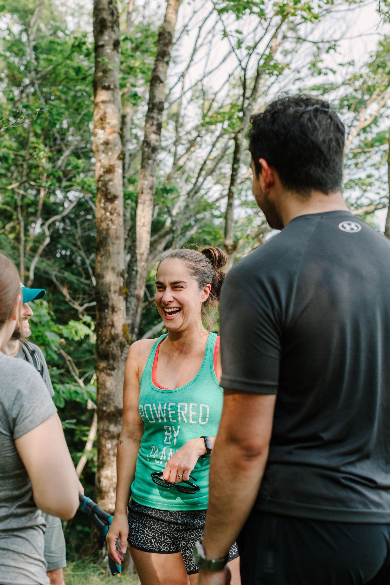 A laughing woman in a green tank top enjoys a conversation with friends outdoors in a wooded setting.