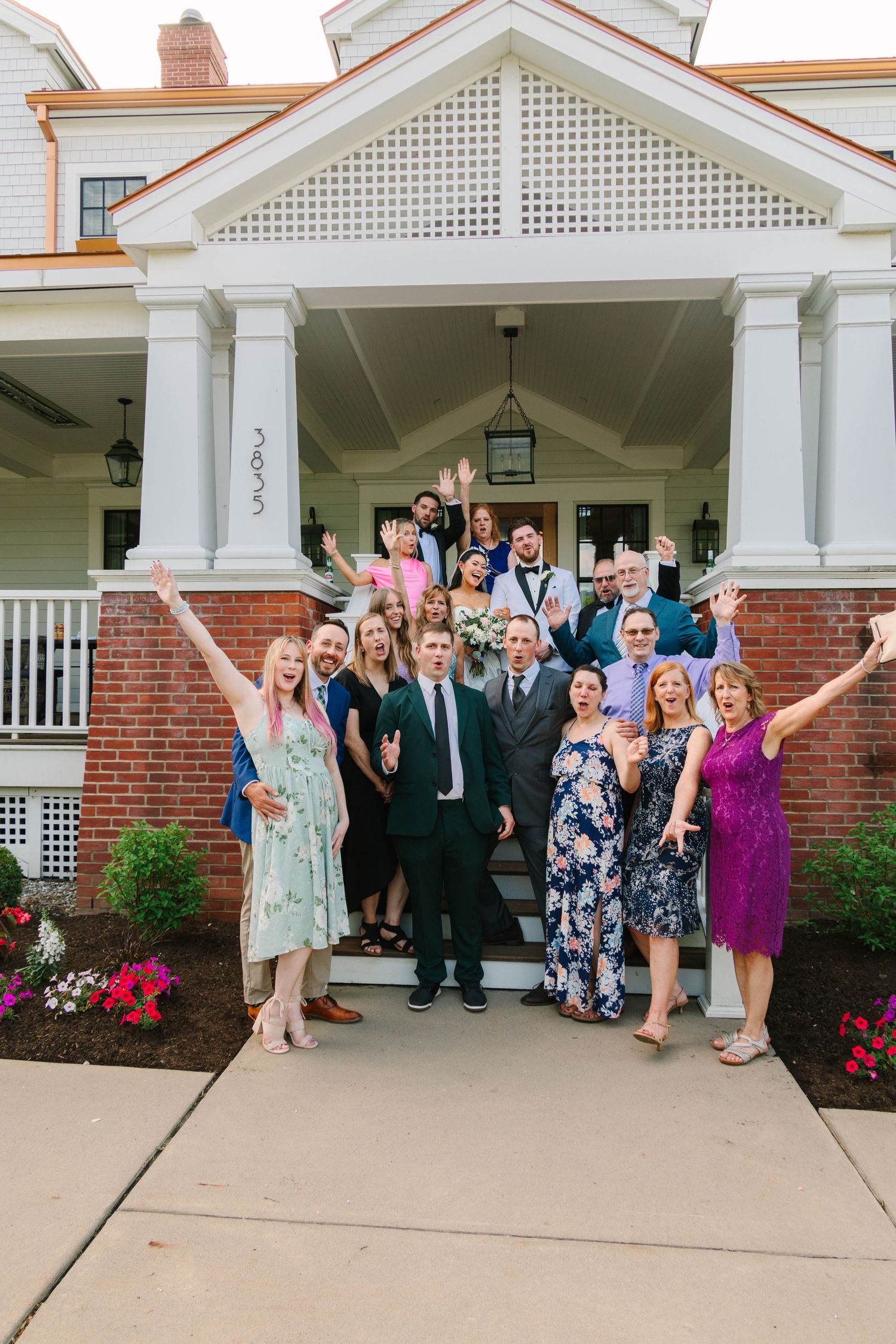 Large wedding group photo on venue steps, guests celebrating with raised arms and joyful expressions.