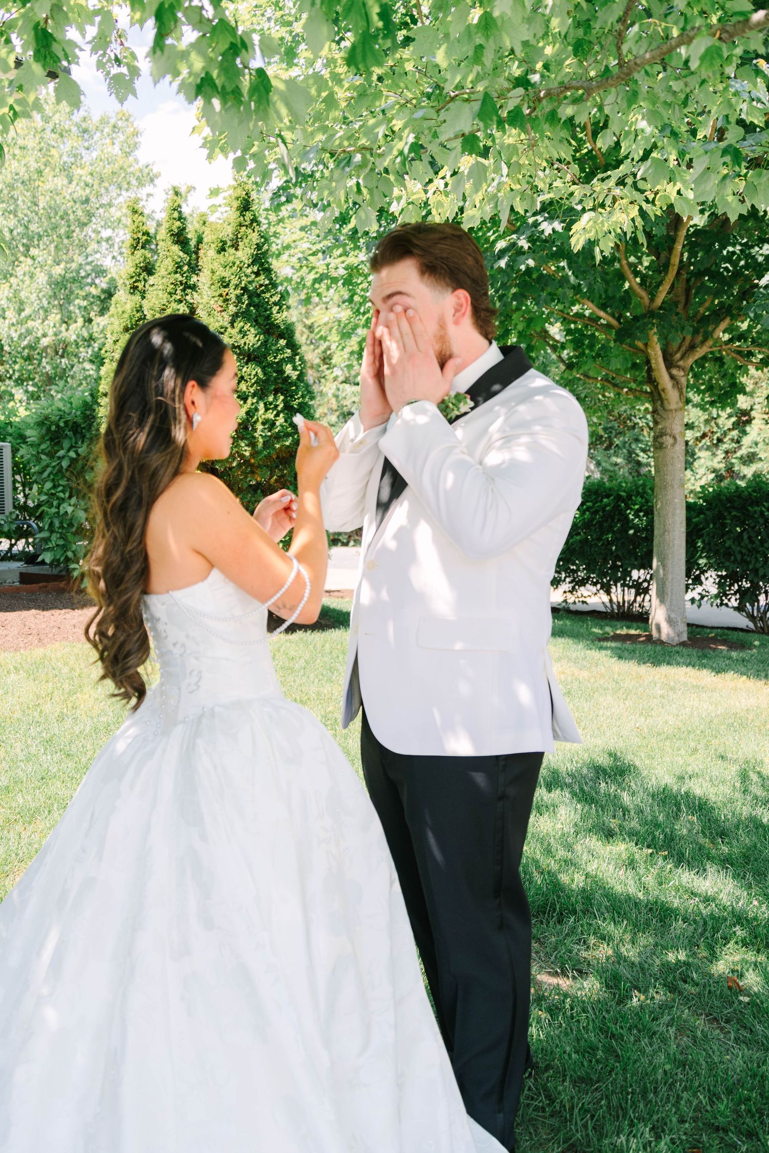 Groom covering his face in emotional reaction during first look with bride in white ballgown.