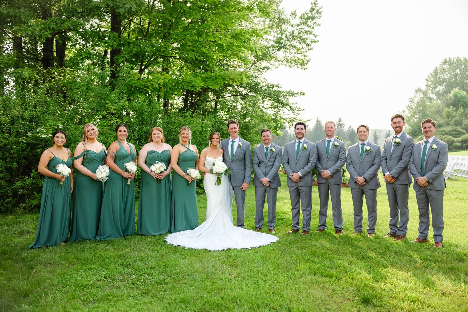 Wedding party posing outdoors with bridesmaids in green gowns and groomsmen in gray suits.