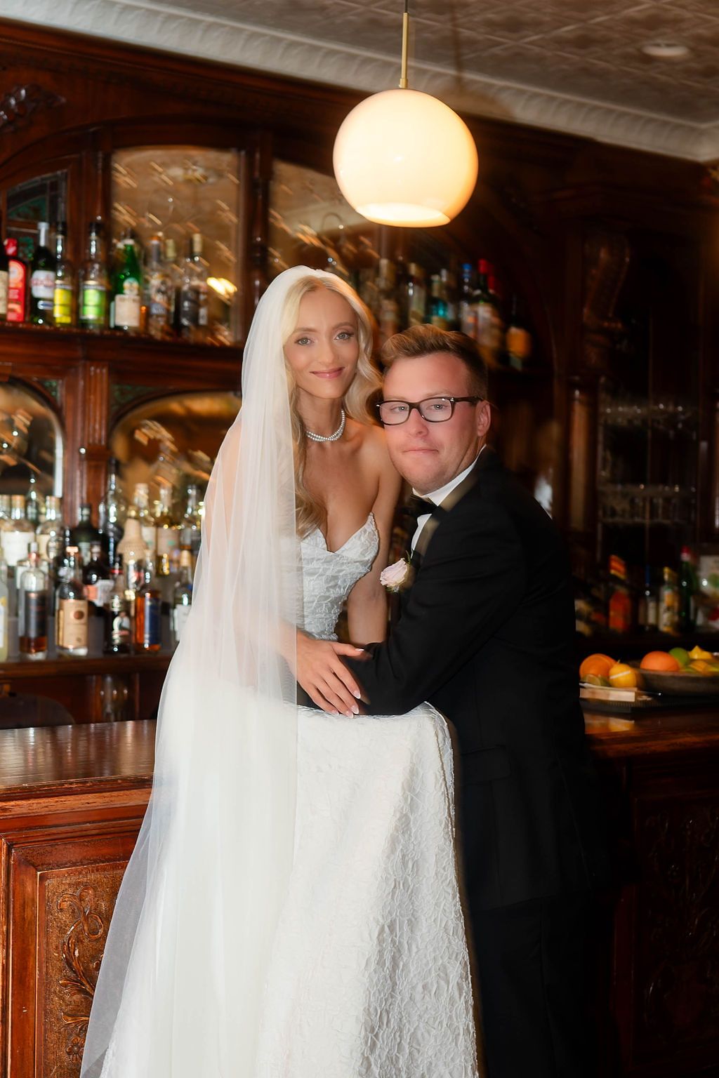 Bride in white lace gown and veil embraces groom in tuxedo at elegant bar venue.