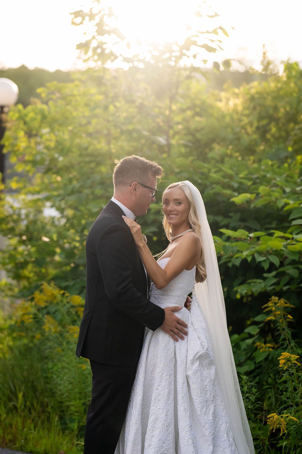Bride and groom embrace in golden sunset light surrounded by lush green garden foliage.