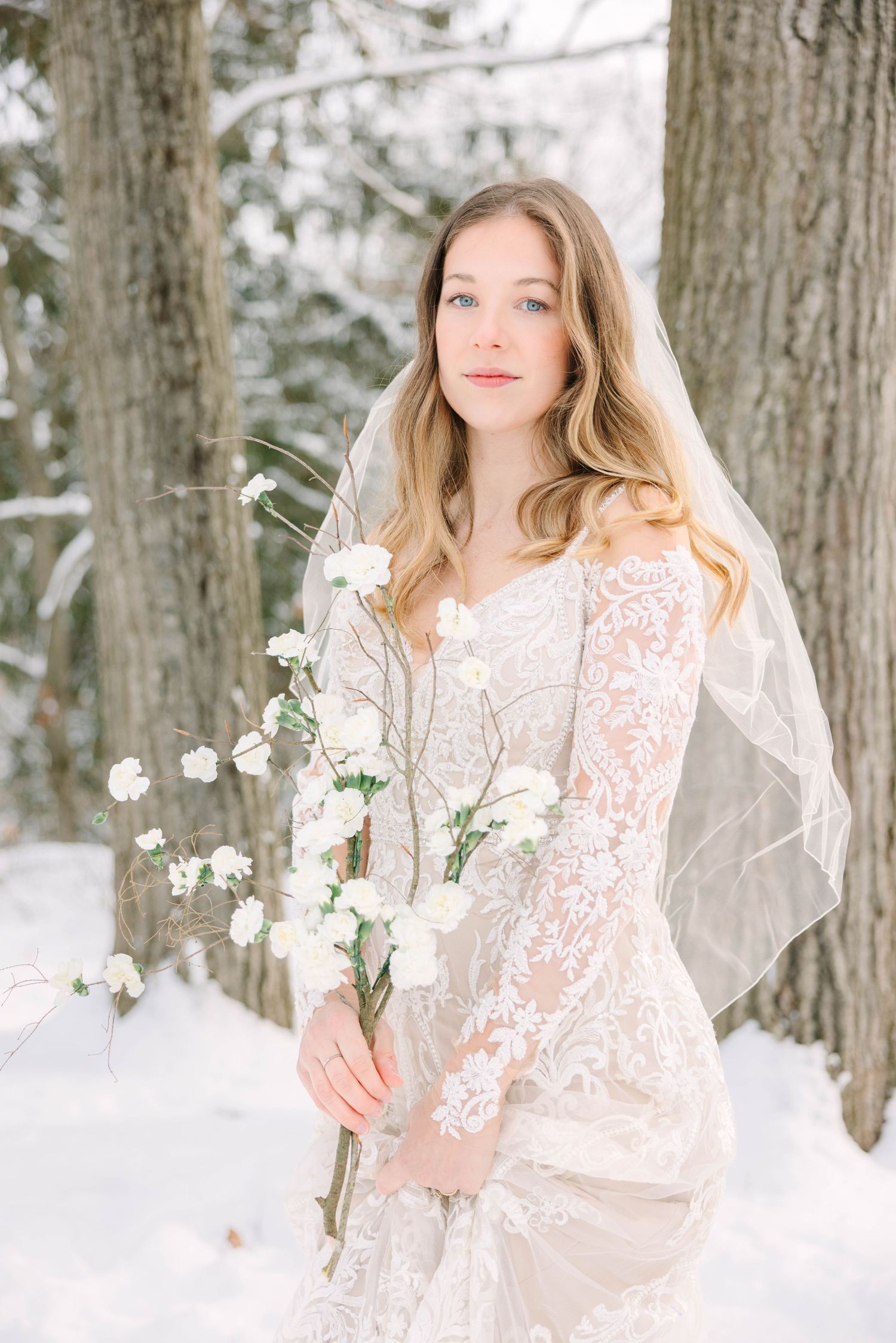 Elegant bride in lace wedding gown and veil holding white flowers in snowy woodland setting.