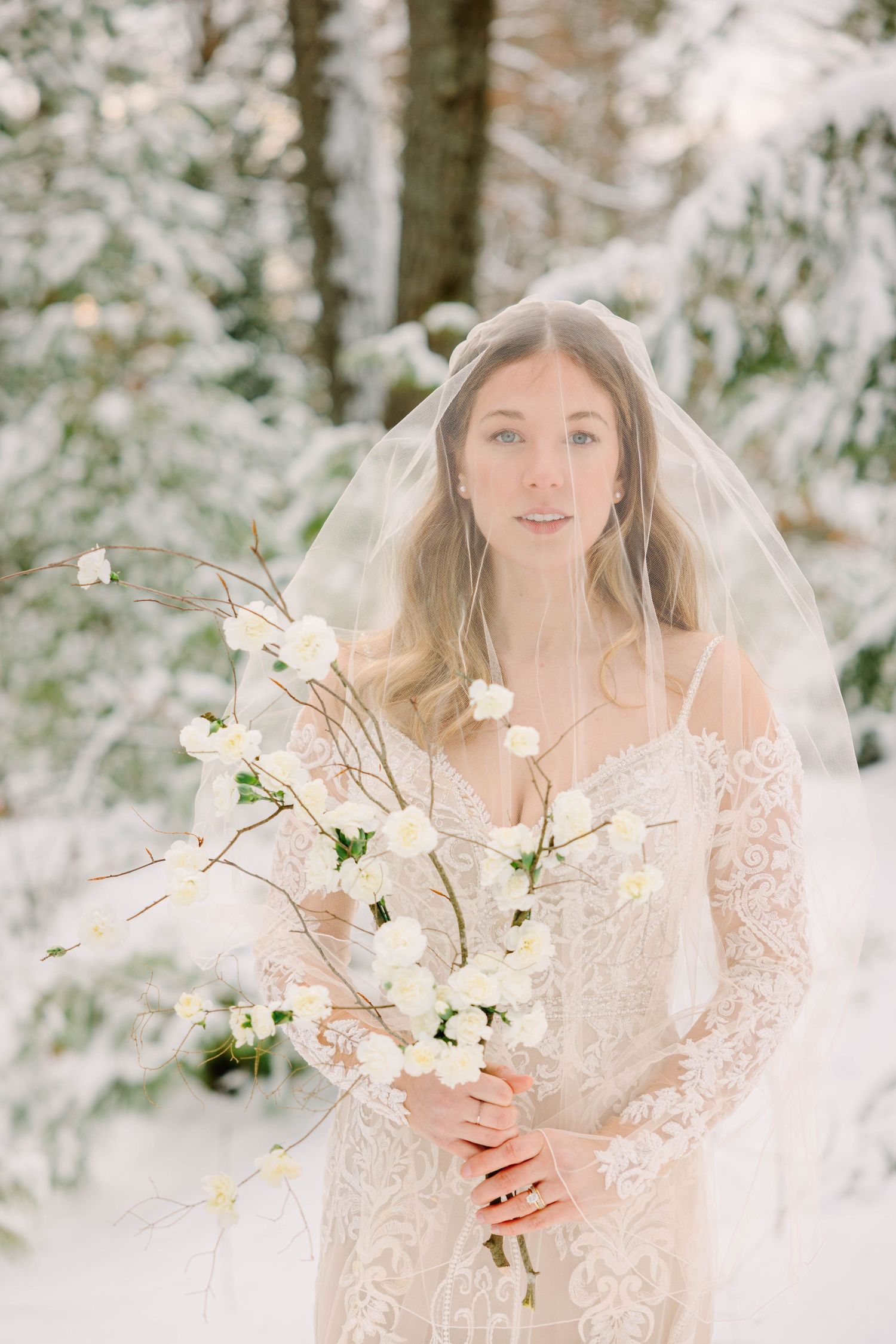 Bride in lace wedding dress and veil holding white flower branch bouquet in snowy forest.