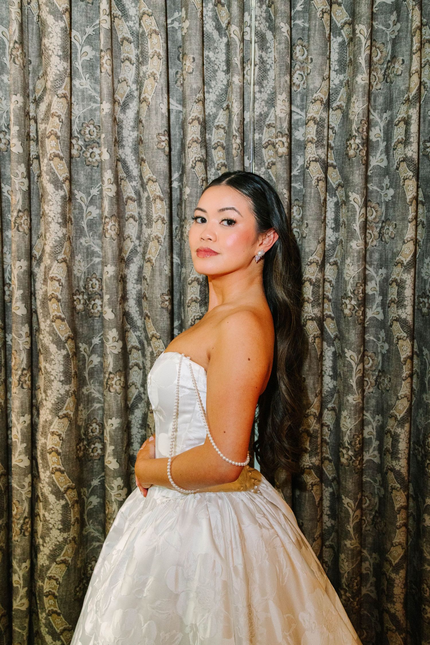 Bride in strapless white ballgown with pearl bracelet posing before patterned curtain backdrop.