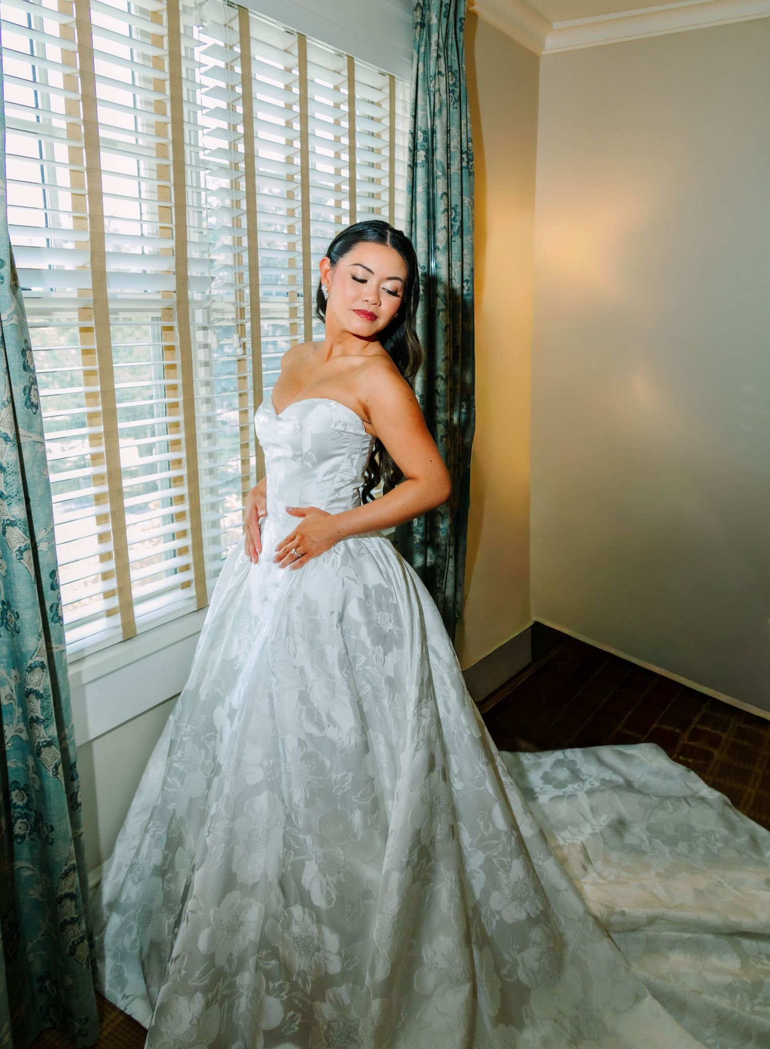 Elegant bride in a strapless floral ballgown wedding dress posing by window with plantation shutters.