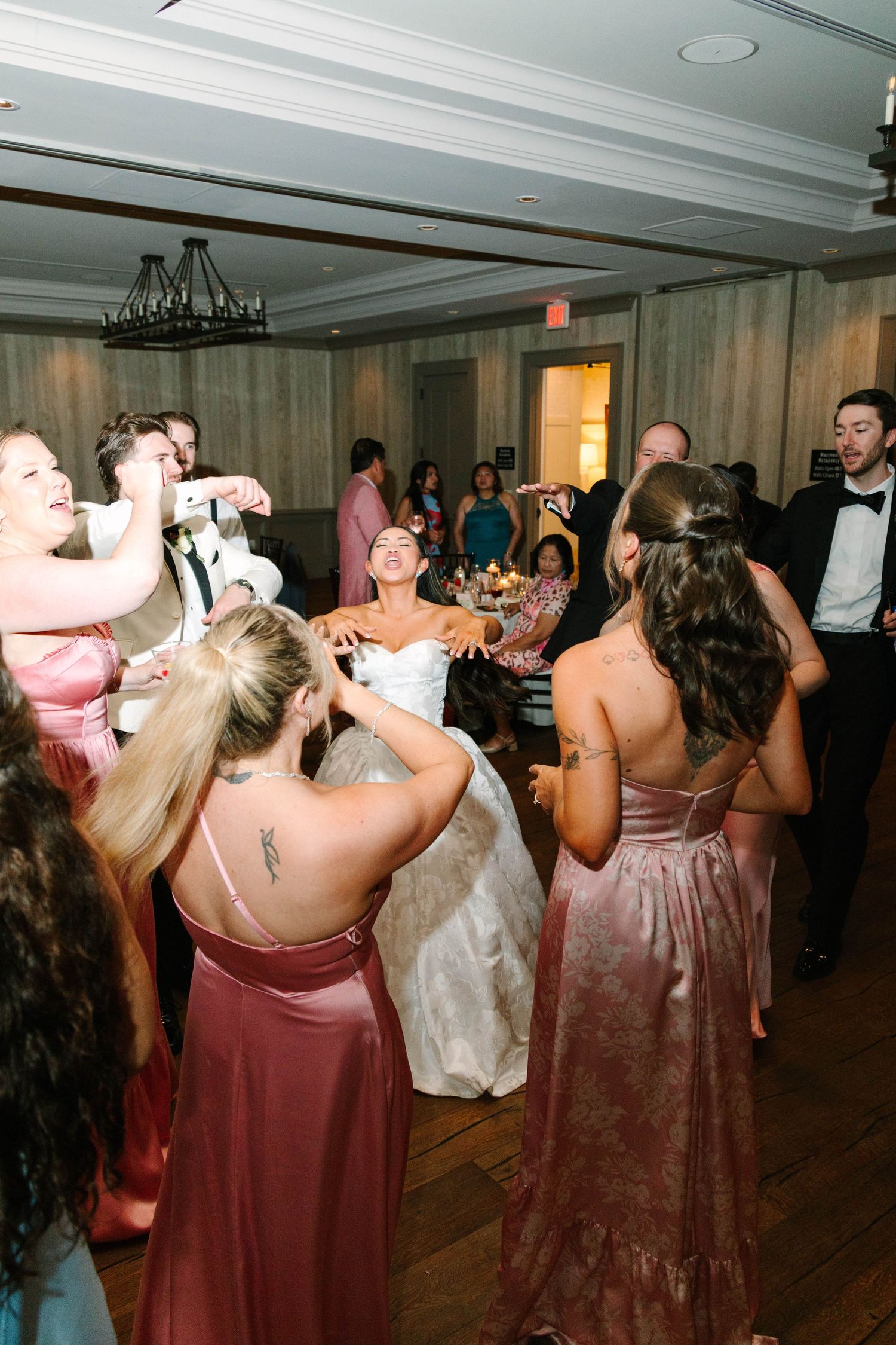 Bride in white gown surrounded by bridesmaids dancing joyfully at wedding reception.