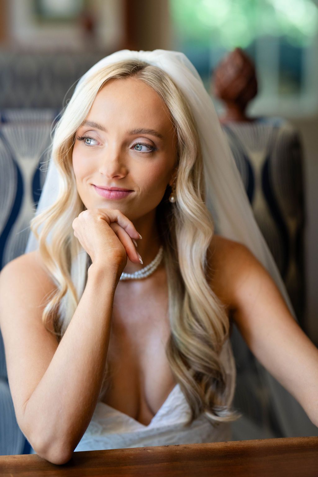 Beautiful blonde bride wearing pearl necklace and veil smiles softly while resting chin on hand.