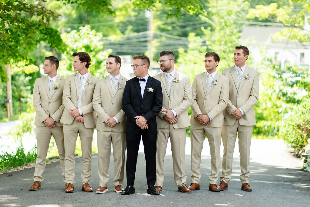 Groom in black tuxedo stands with groomsmen dressed in matching tan suits outdoors on a sunny day.