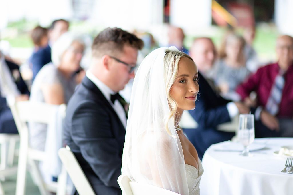 Radiant bride in white veil smiling during wedding reception speeches inside a bright elegant tent venue.