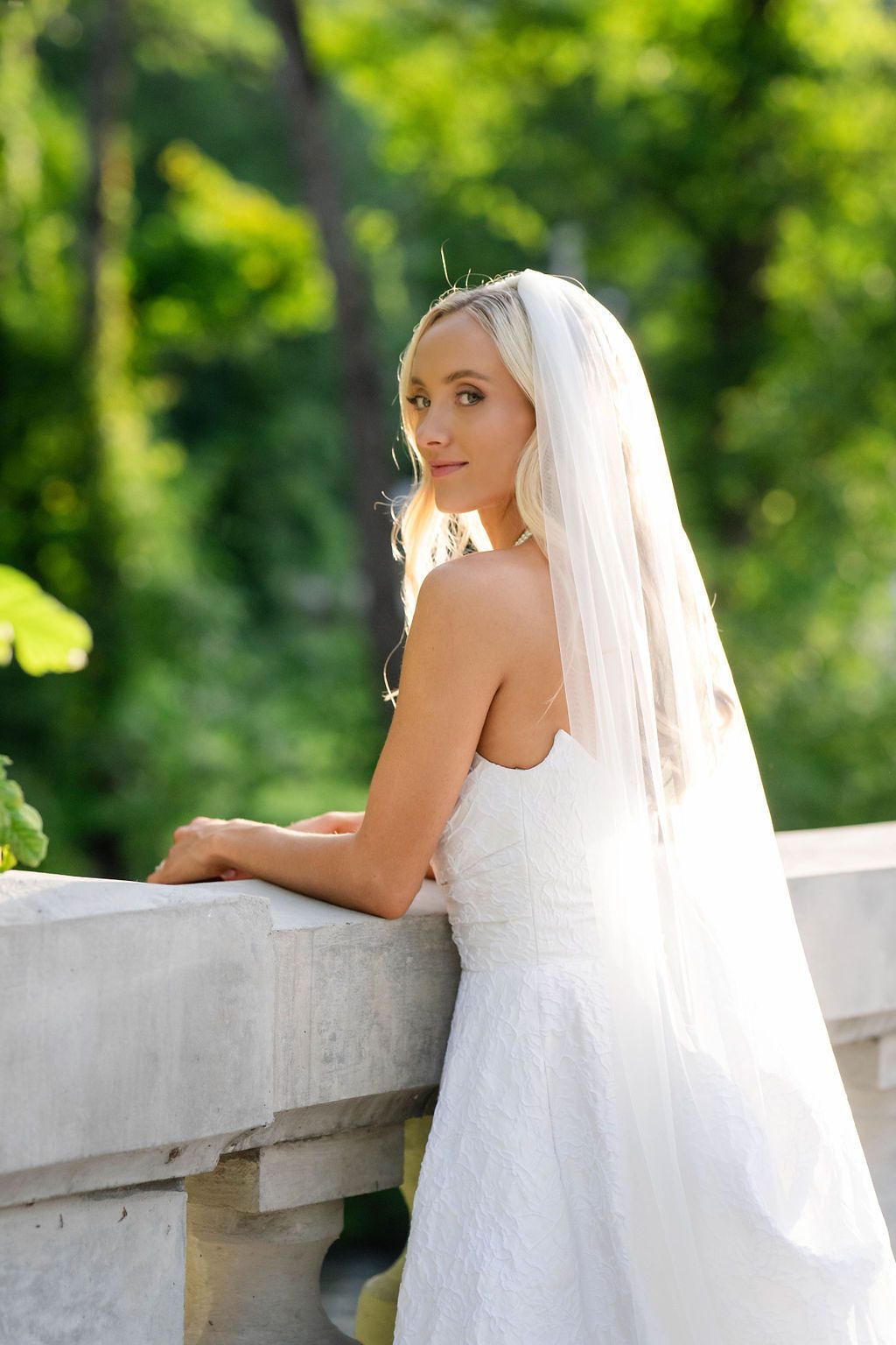 Bride in white strapless lace gown and veil looking over shoulder while leaning on stone balustrade outdoors.