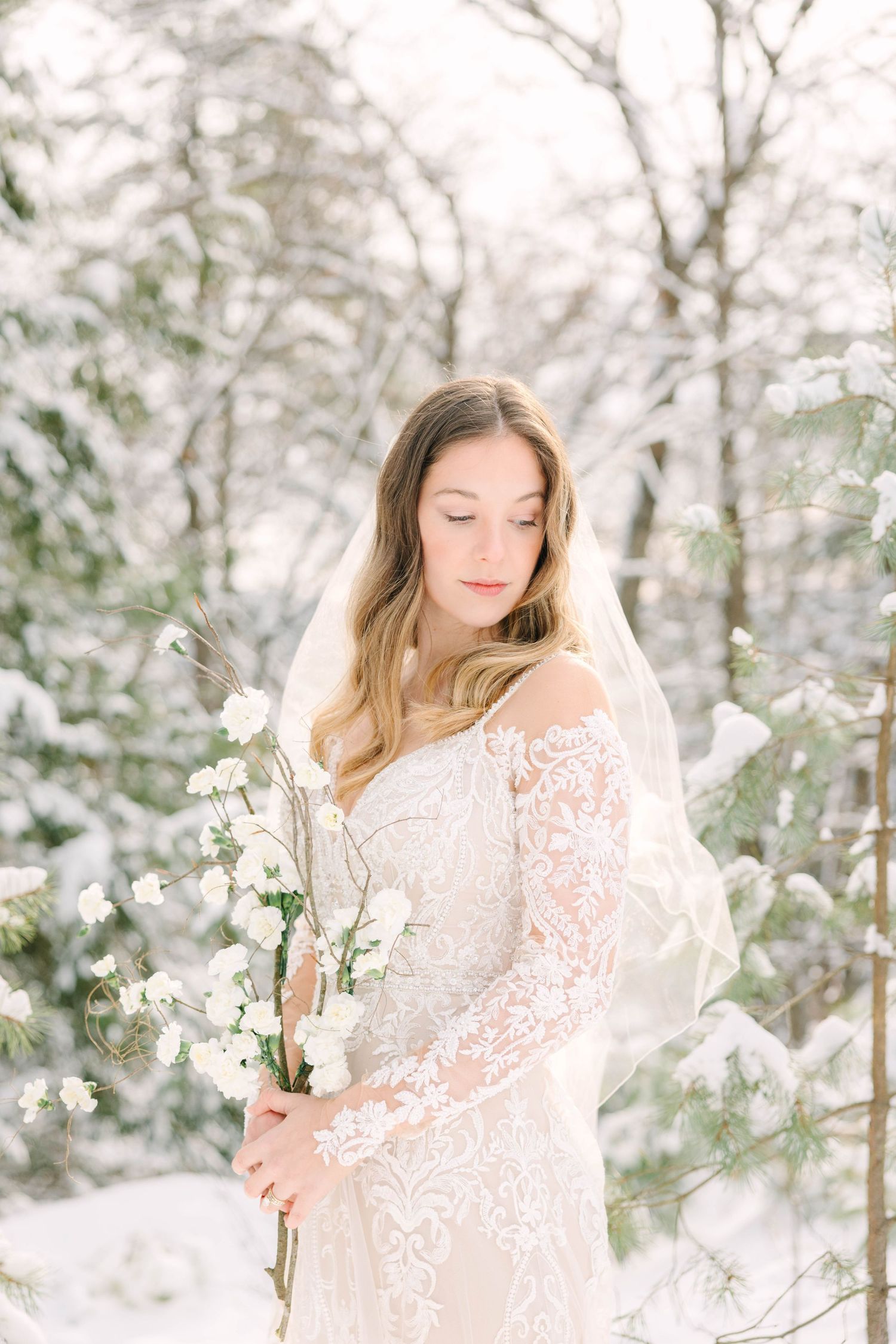 Bride in lace off-shoulder gown and veil holds white flowers in snowy winter forest.