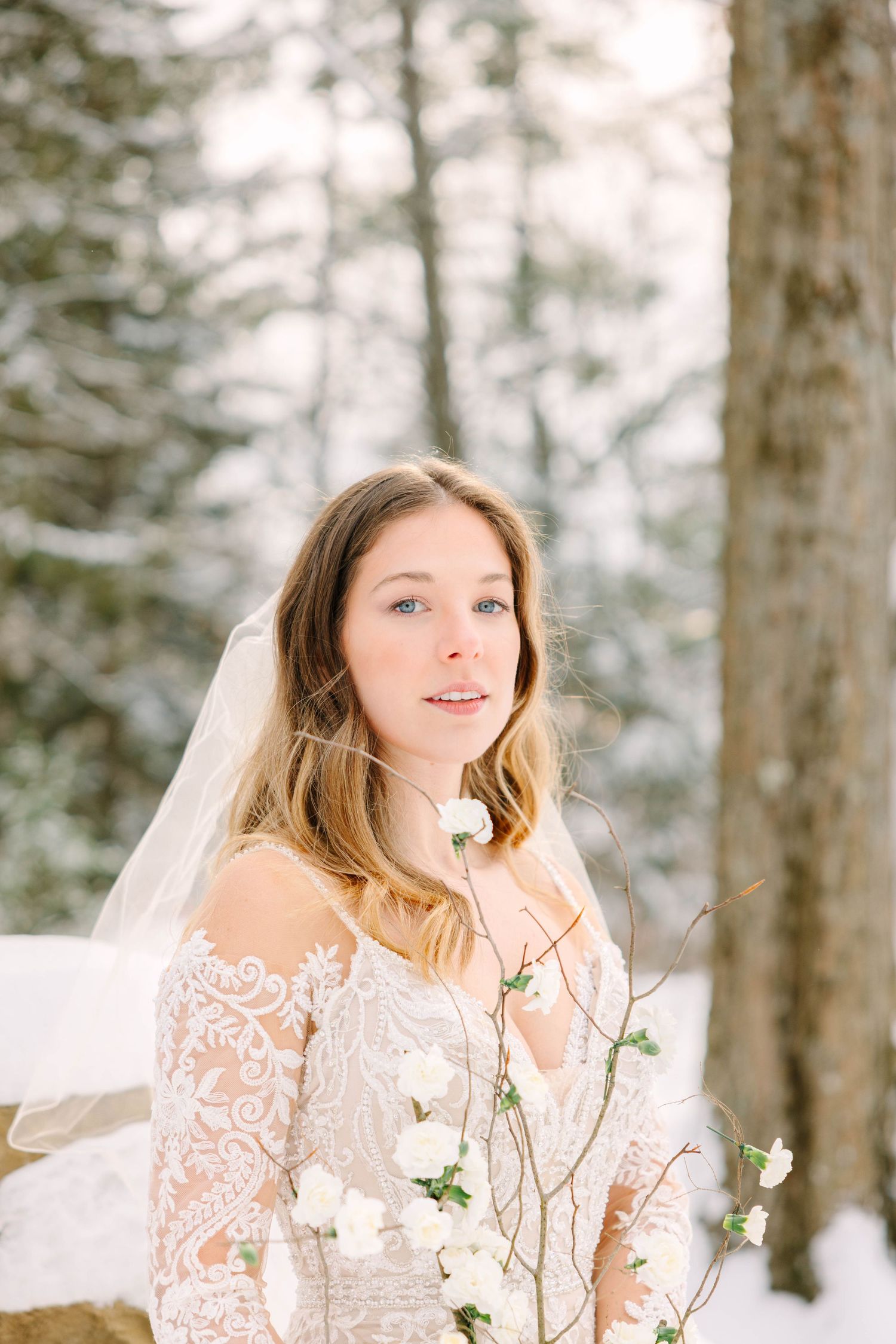 Portrait of a bride in lace gown and veil holding white floral branch in a bright snowy winter forest.