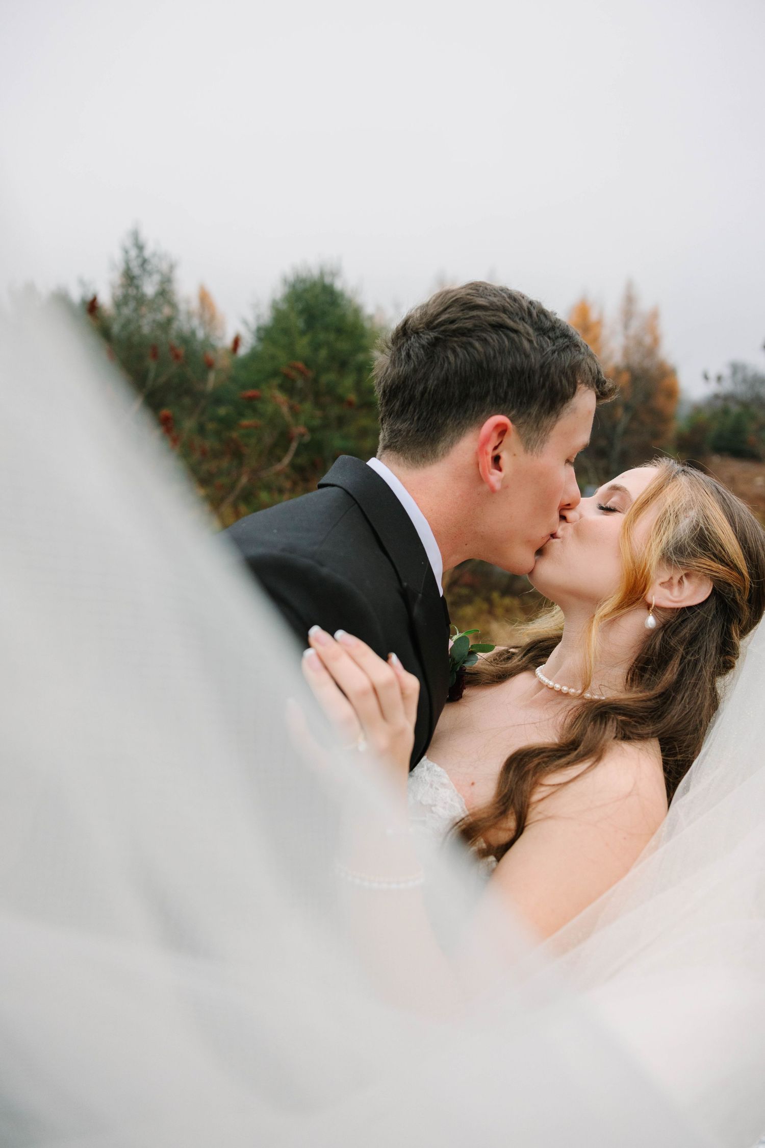 Bride and groom sharing romantic kiss outdoors with veil flowing in foreground against autumn trees.