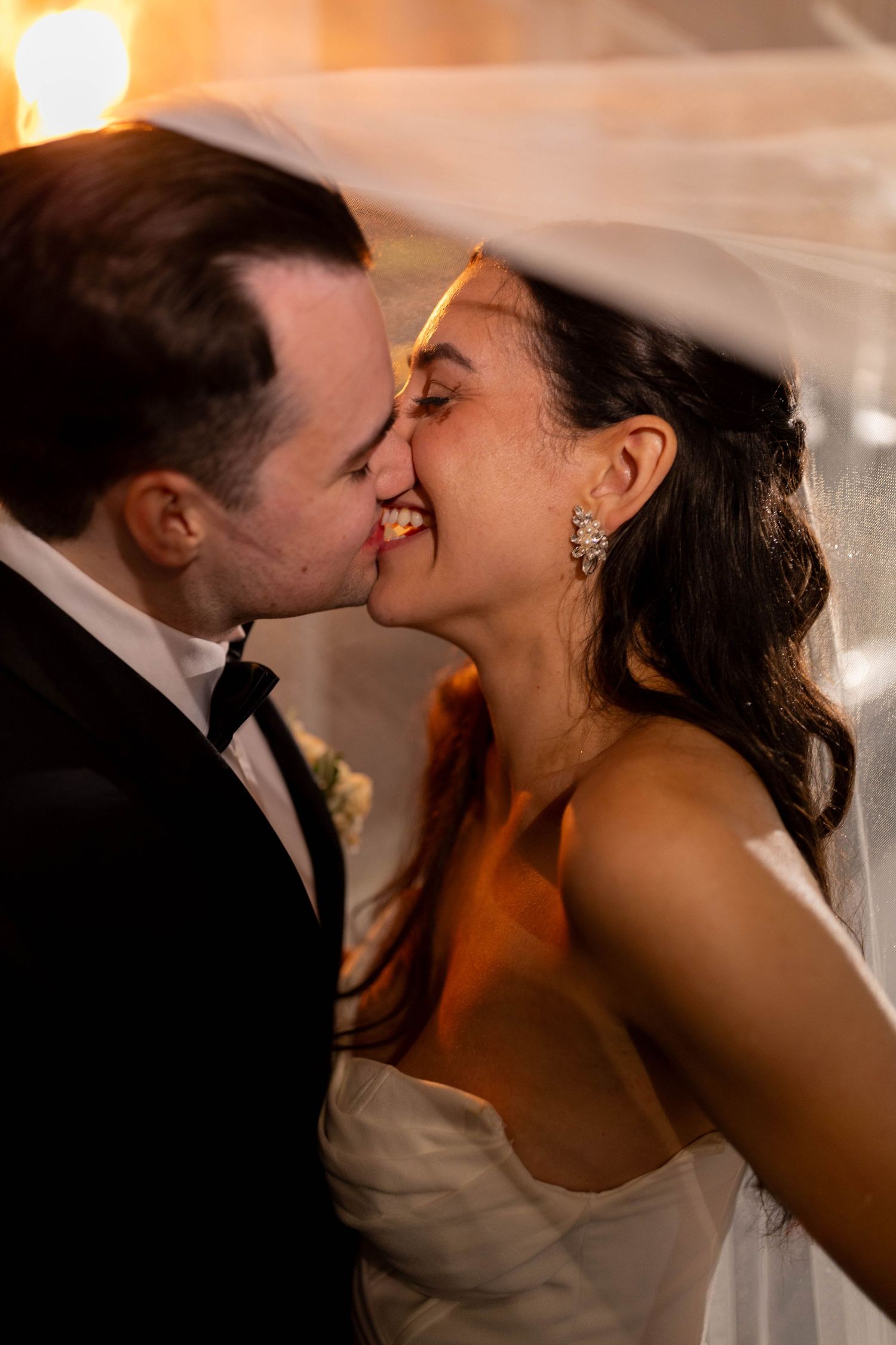 Bride and groom share an intimate moment under veil with warm golden bokeh lighting.