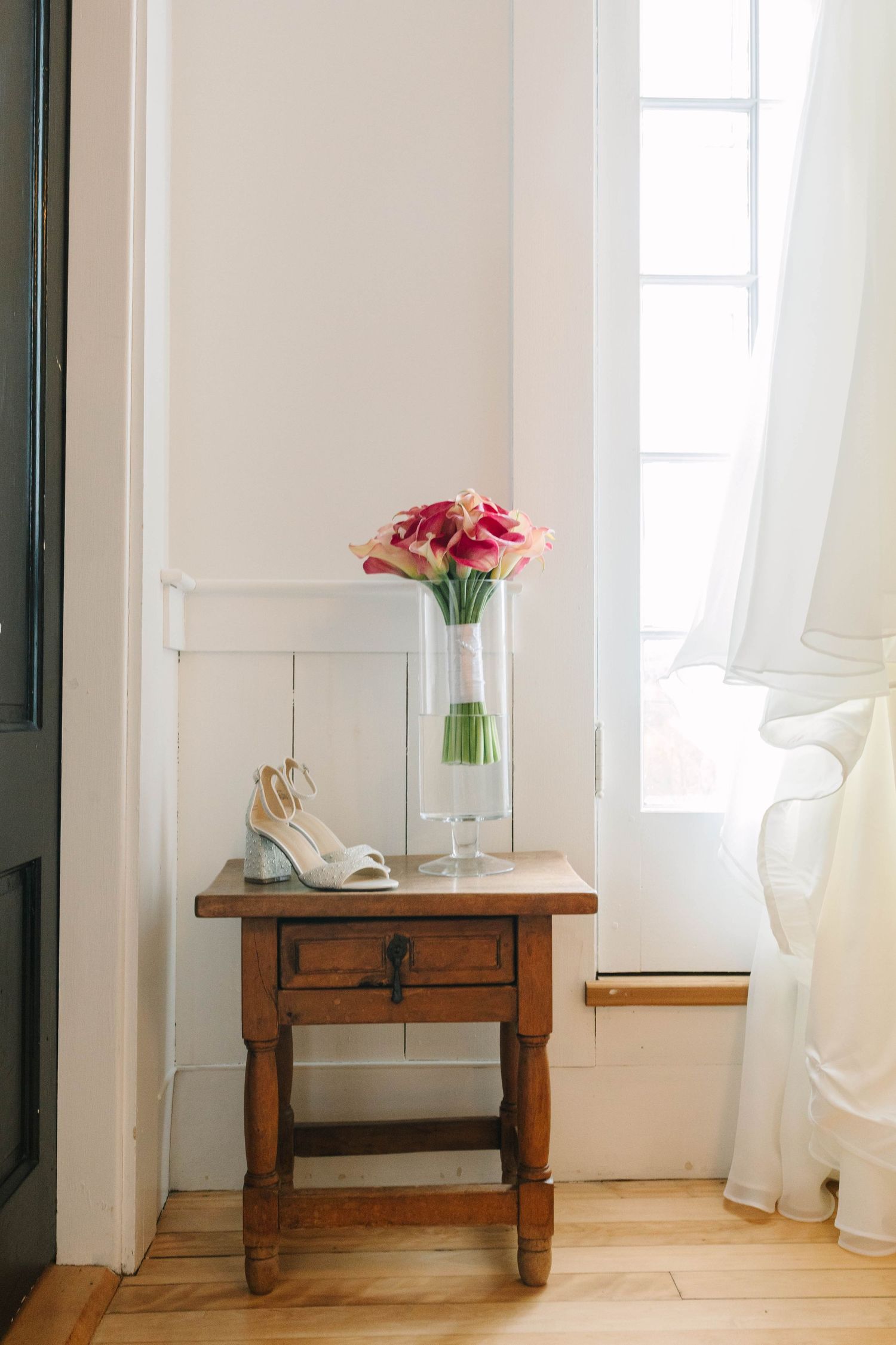Pink calla lily bouquet in glass vase with white bridal heels on rustic wooden side table near bright window.