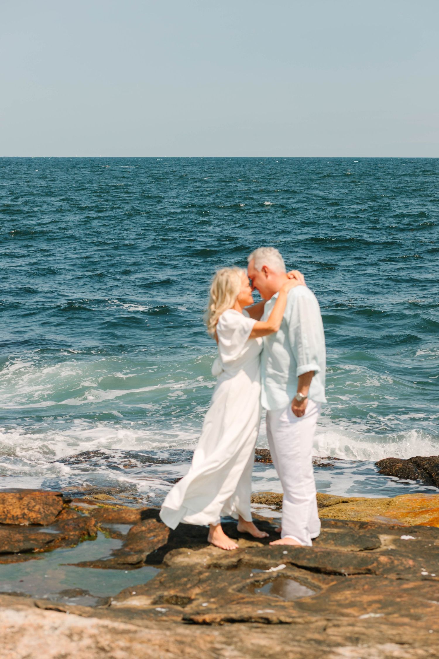 Romantic senior couple in white clothing kissing on rocky ocean shore with blue sea waves in background.