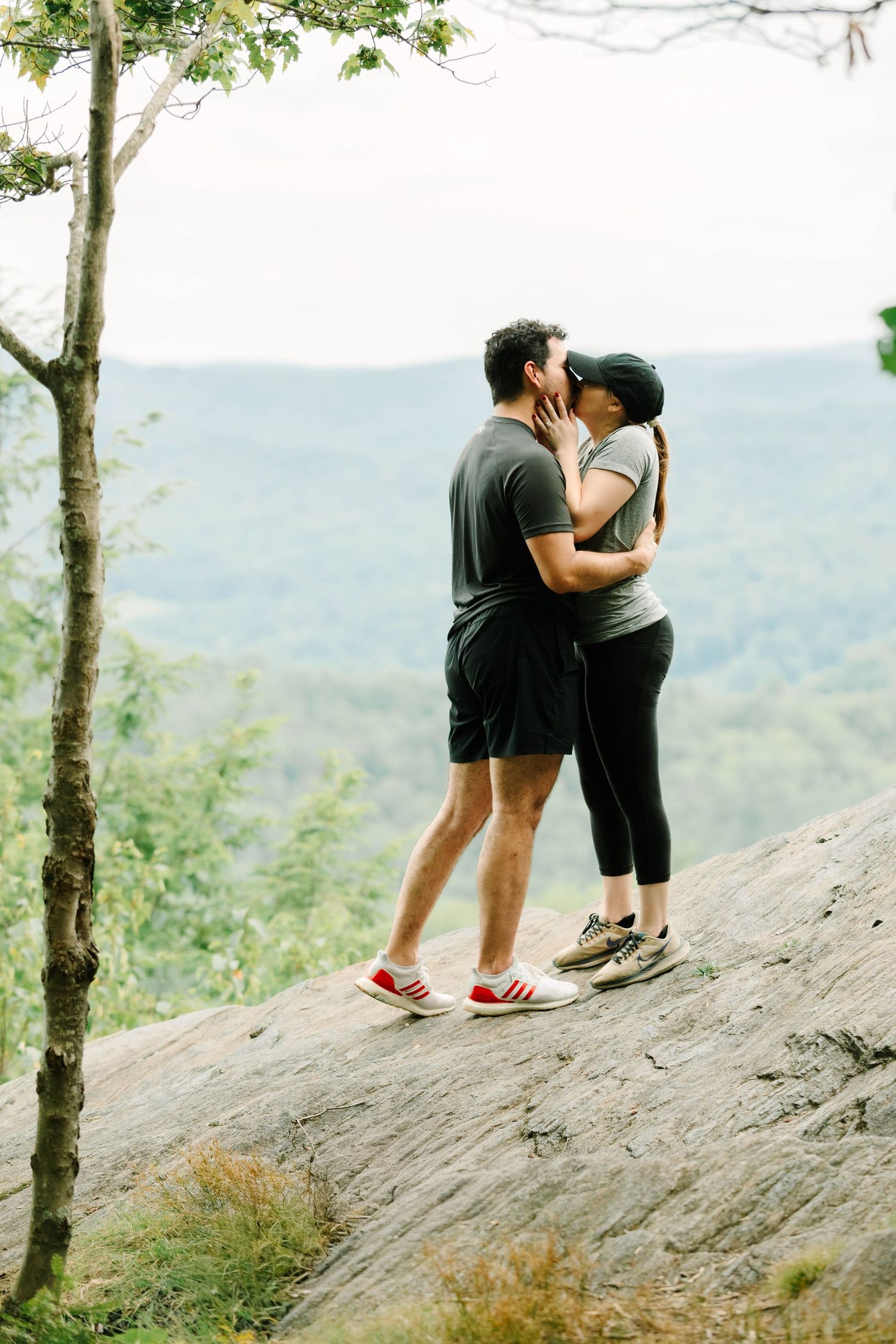 Couple kissing on a rocky overlook with lush green forest valley views behind them.