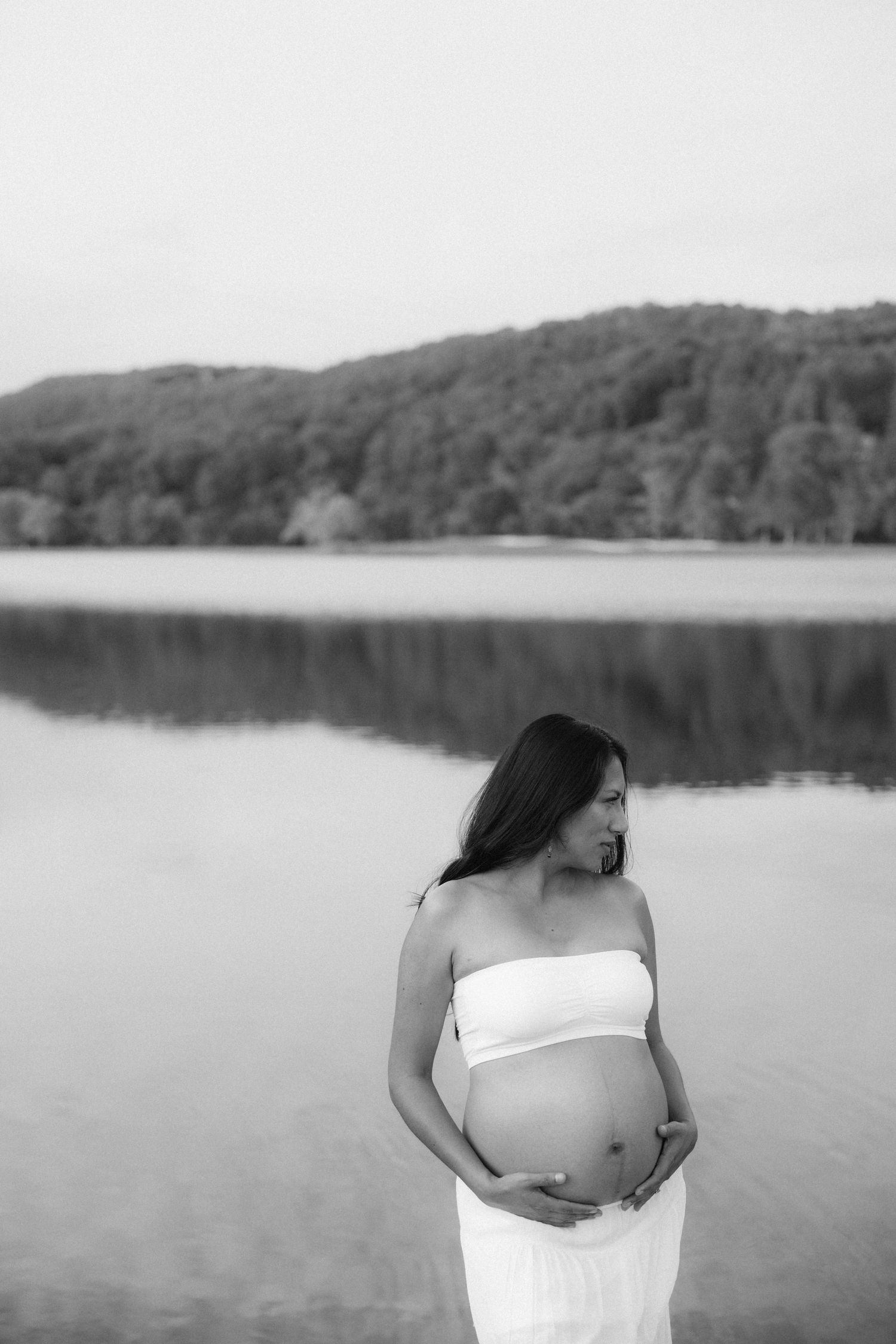 Black and white photo of a pregnant woman in a white dress standing by a serene lake with forested hills in the background.