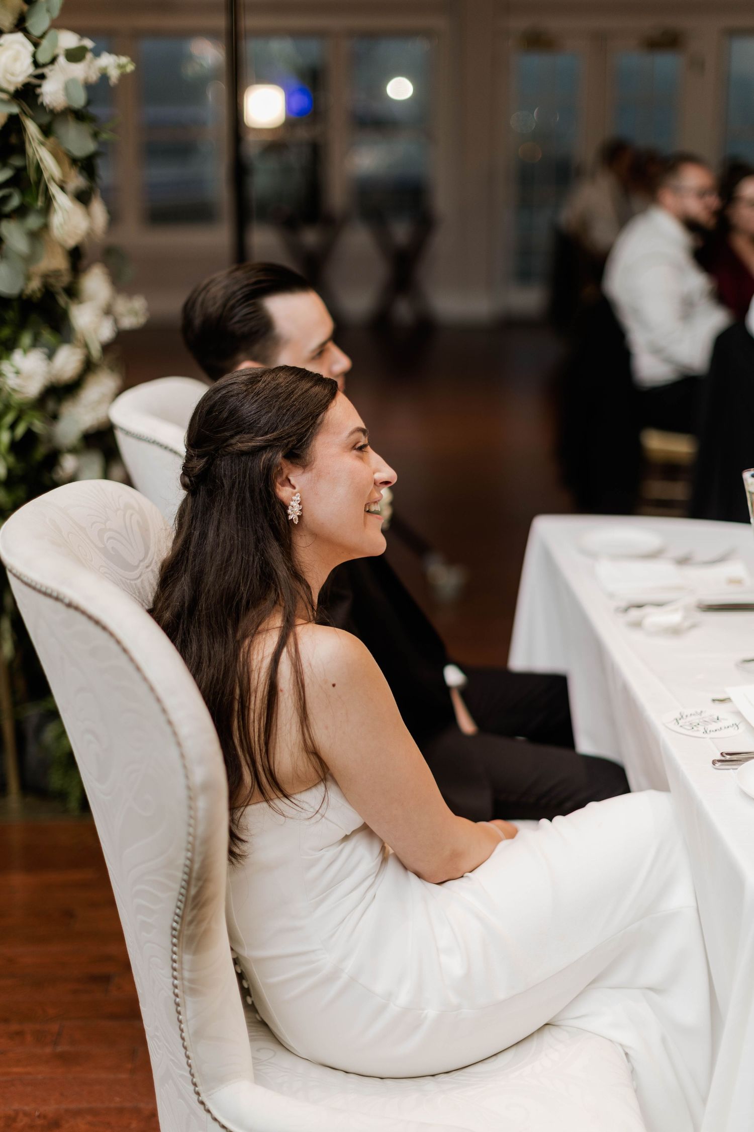 Bride in white strapless gown smiles while seated in elegant white chair at wedding reception dinner table.