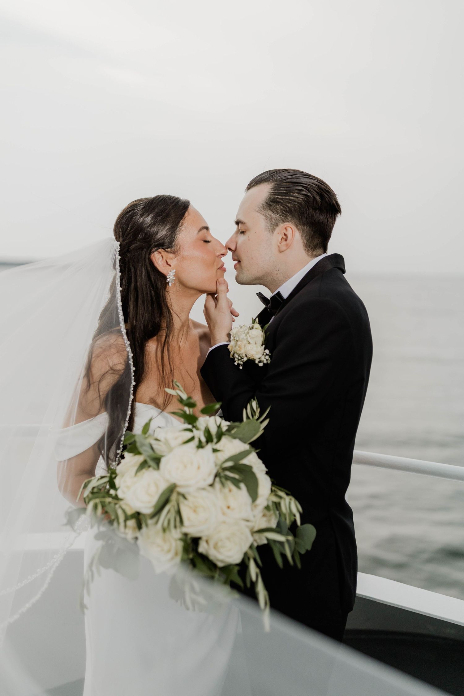 Bride and groom share romantic kiss on boat deck, bride holding white rose bouquet with flowing veil.