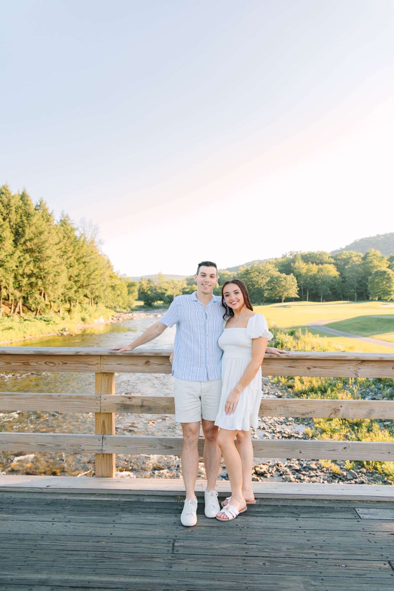 Happy couple posing together on a wooden bridge overlooking a scenic river valley at golden hour.