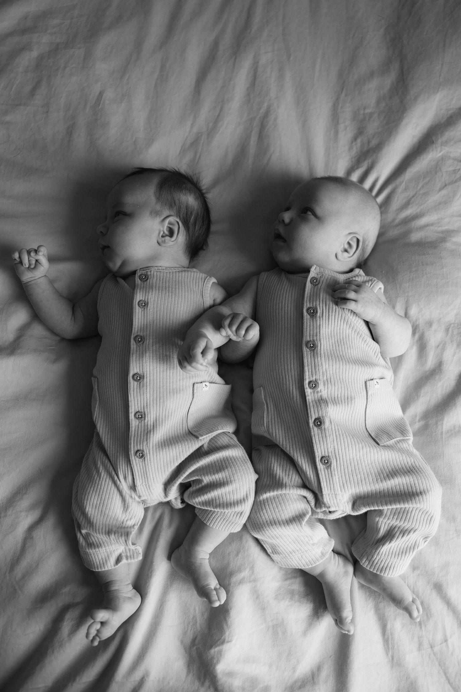 Black and white overhead photo of newborn twins in matching button rompers lying on a bed.