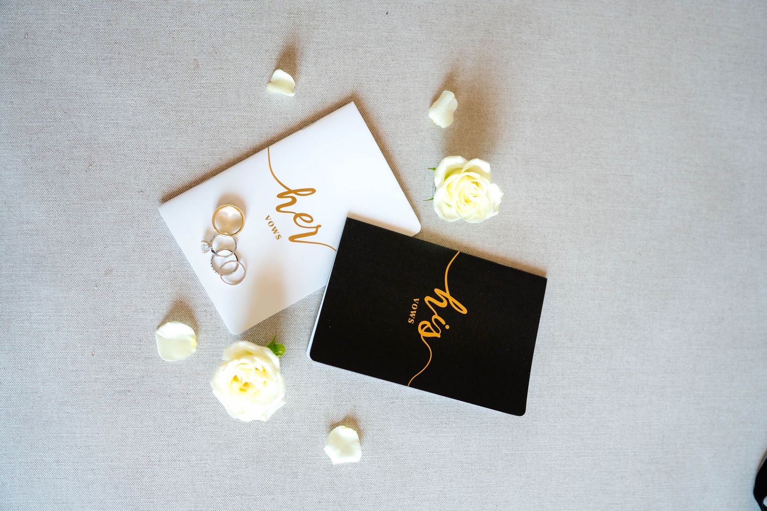Flat lay of black and white wedding vow books with gold script, rings, and white roses on linen.