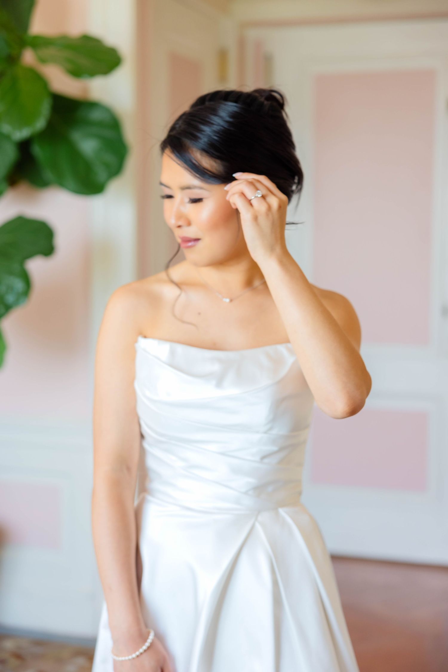 Bride in strapless white satin gown adjusting her hair, wearing pearl bracelet and diamond ring.