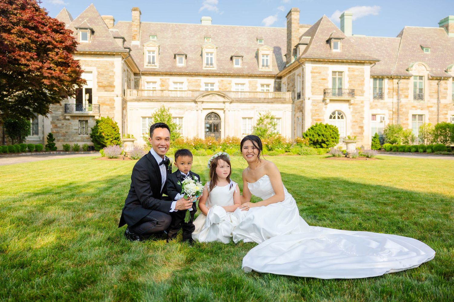 Wedding couple with two children posing on lawn in front of elegant stone manor house.